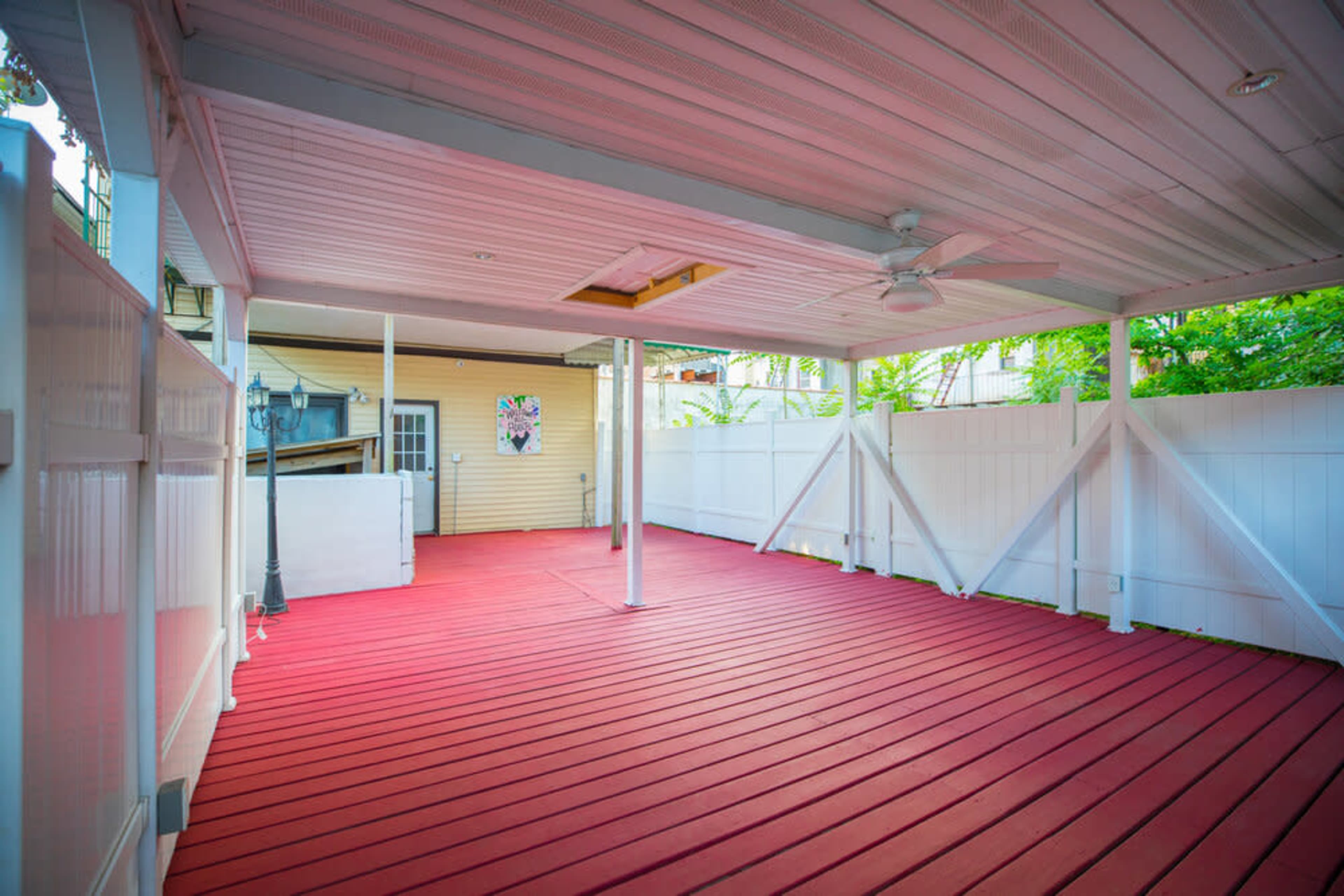 A covered outdoor patio with red wooden decking and white fencing, featuring a ceiling fan overhead.