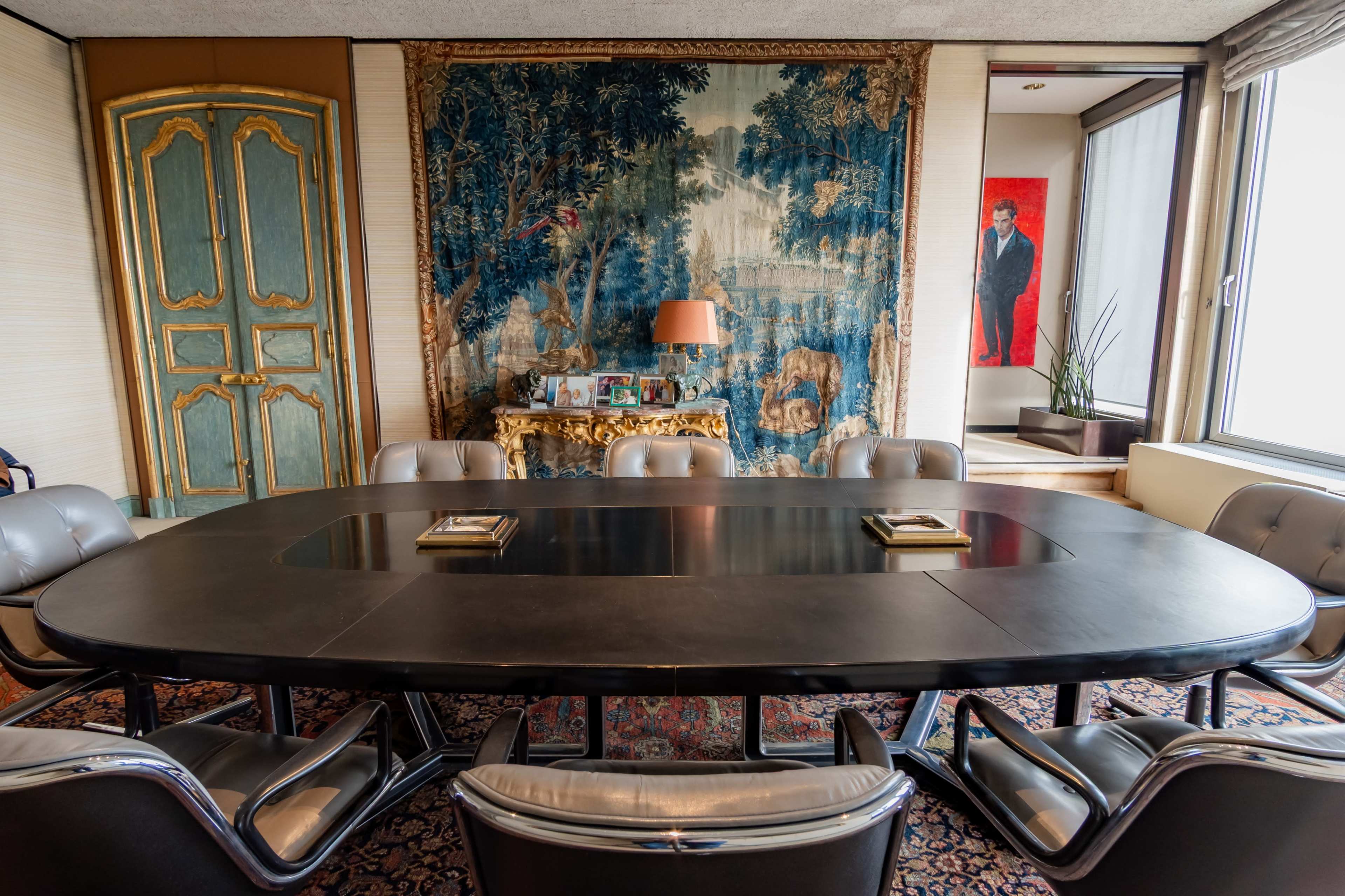 The image shows a conference room with a large oval table surrounded by modern chairs, adorned with a tapestry on one wall and artwork on another.