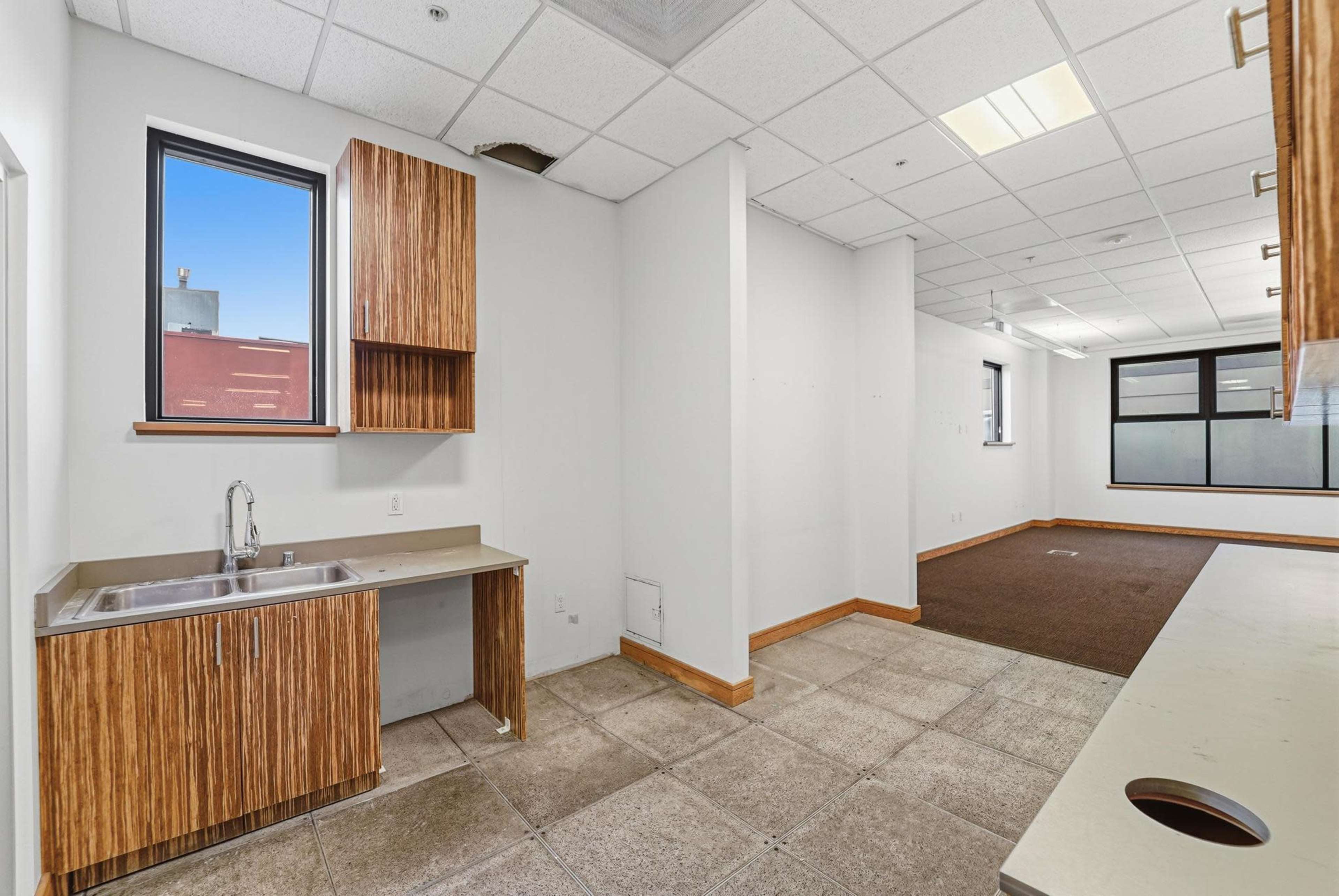 The image shows an empty commercial kitchen area with a stainless steel sink and wooden cabinetry, adjacent to a bare open space with carpeted flooring.
