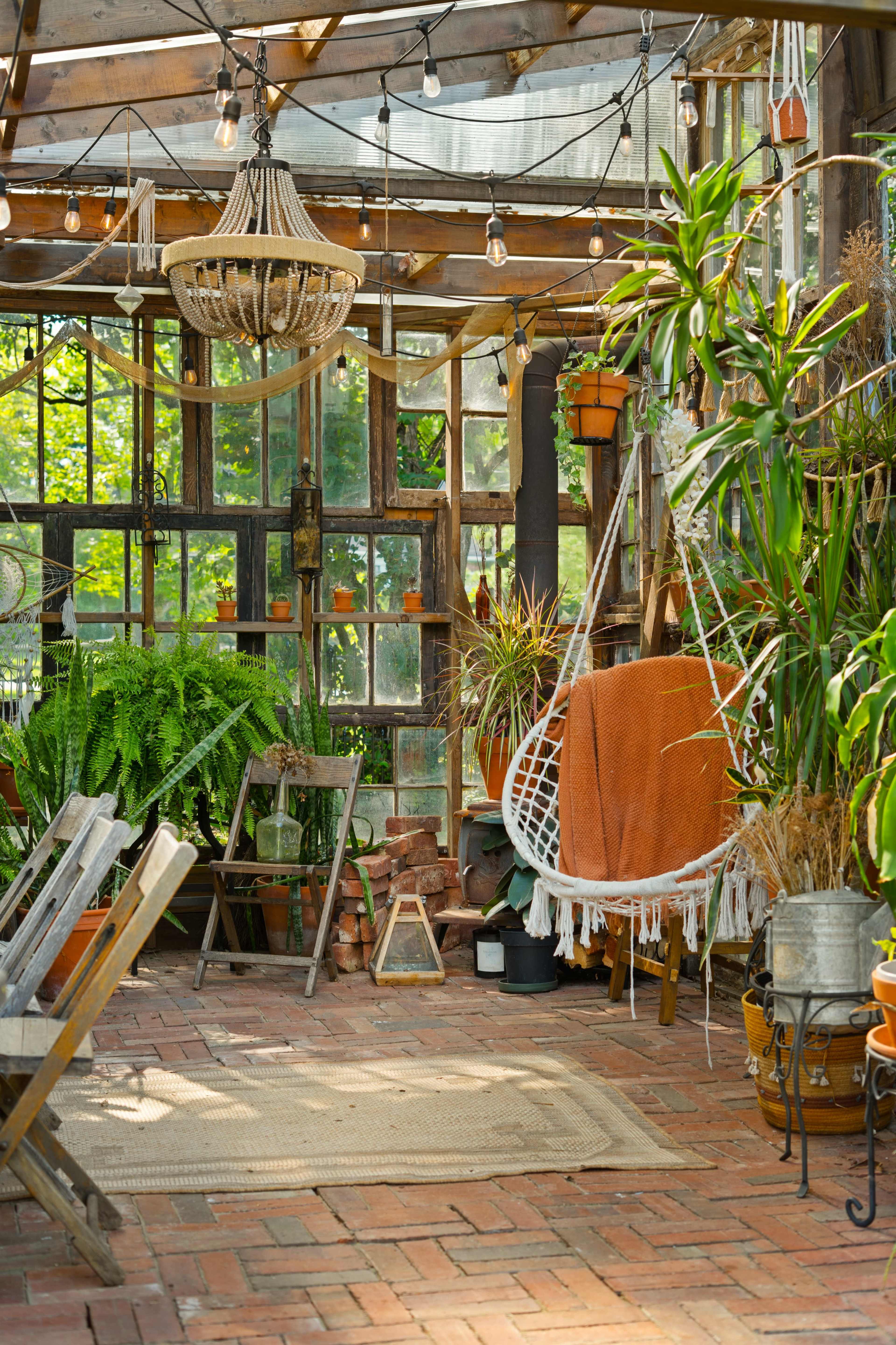 The image depicts a cozy greenhouse interior with hanging plants, a hammock, and rustic furniture arranged on brick flooring.