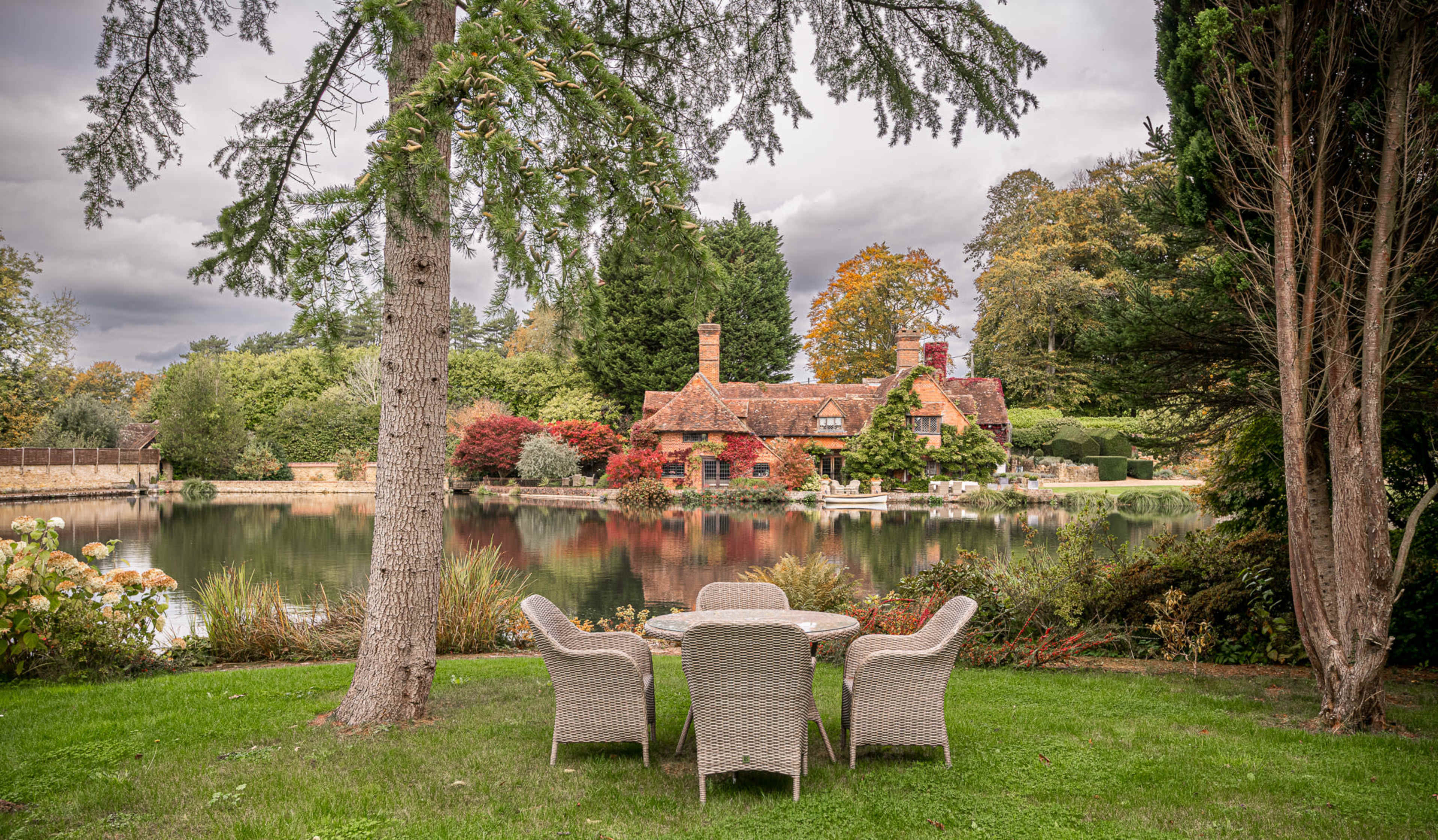 A cottage with red and brown foliage is situated by a calm lake, surrounded by a manicured garden and outdoor furniture.