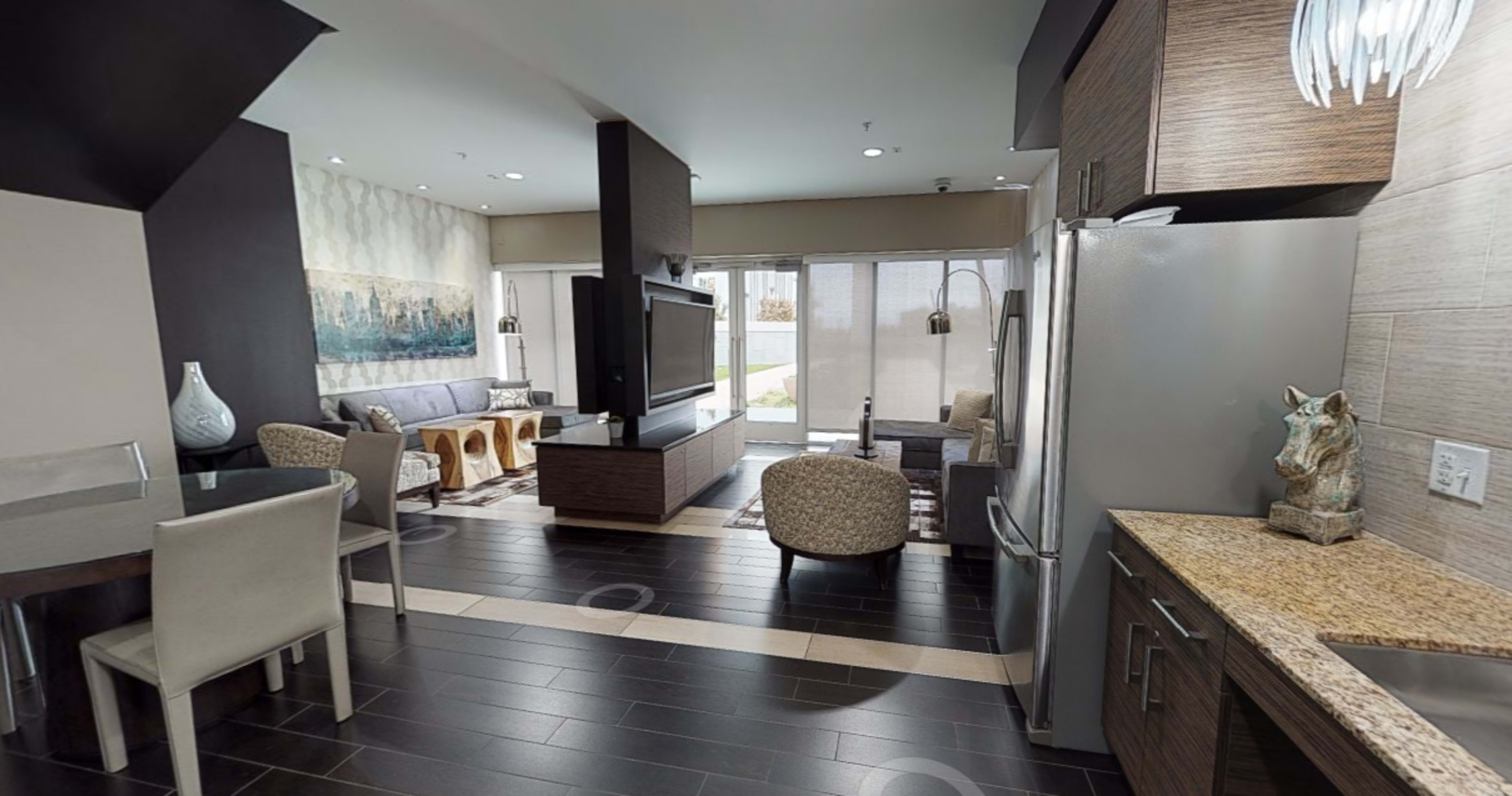 The image shows a modern open-concept living space with a kitchen, dining area, and living room featuring large windows and contemporary furniture.