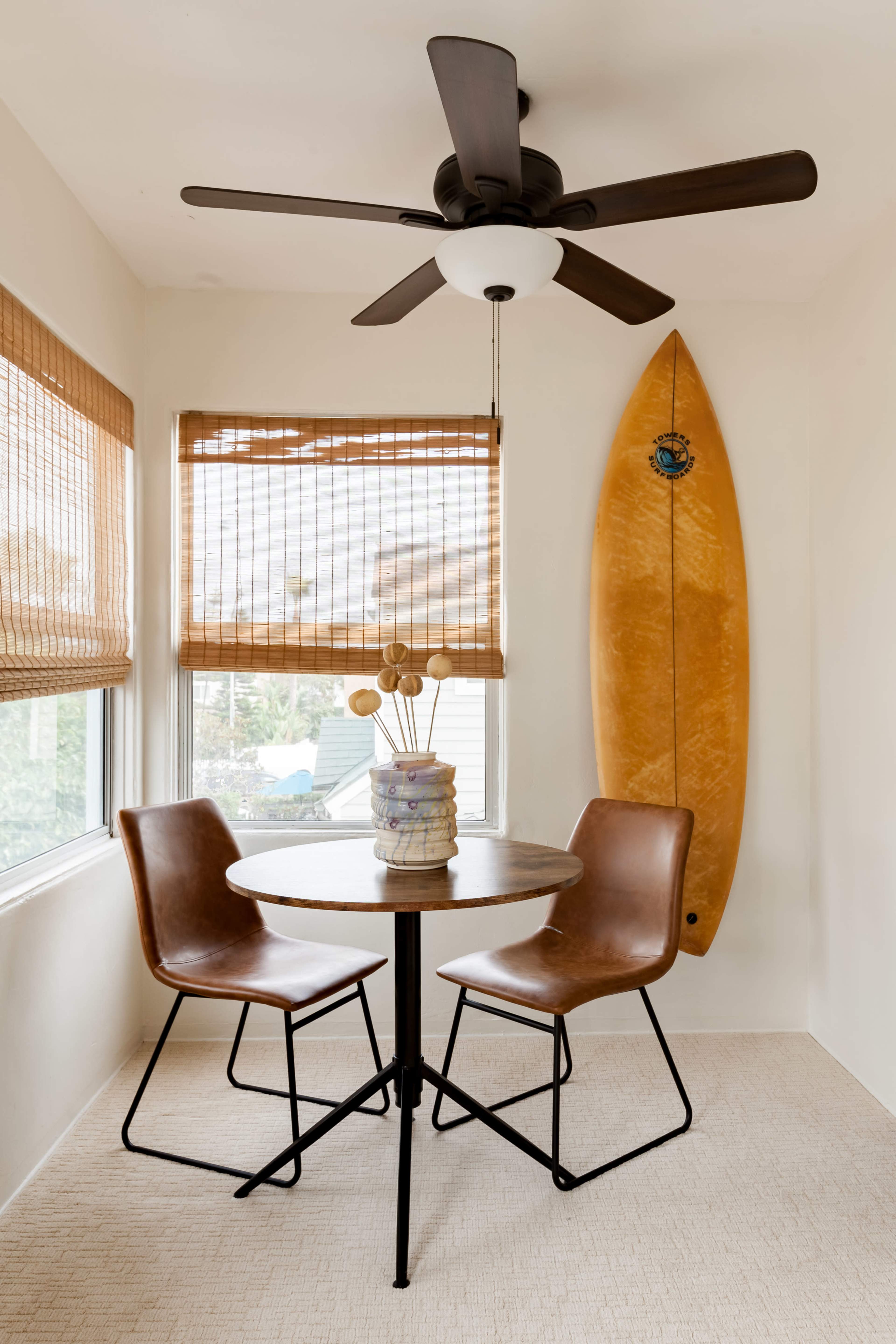 A small dining area features a round table with two brown chairs, a ceiling fan, and a surfboard mounted on the wall.