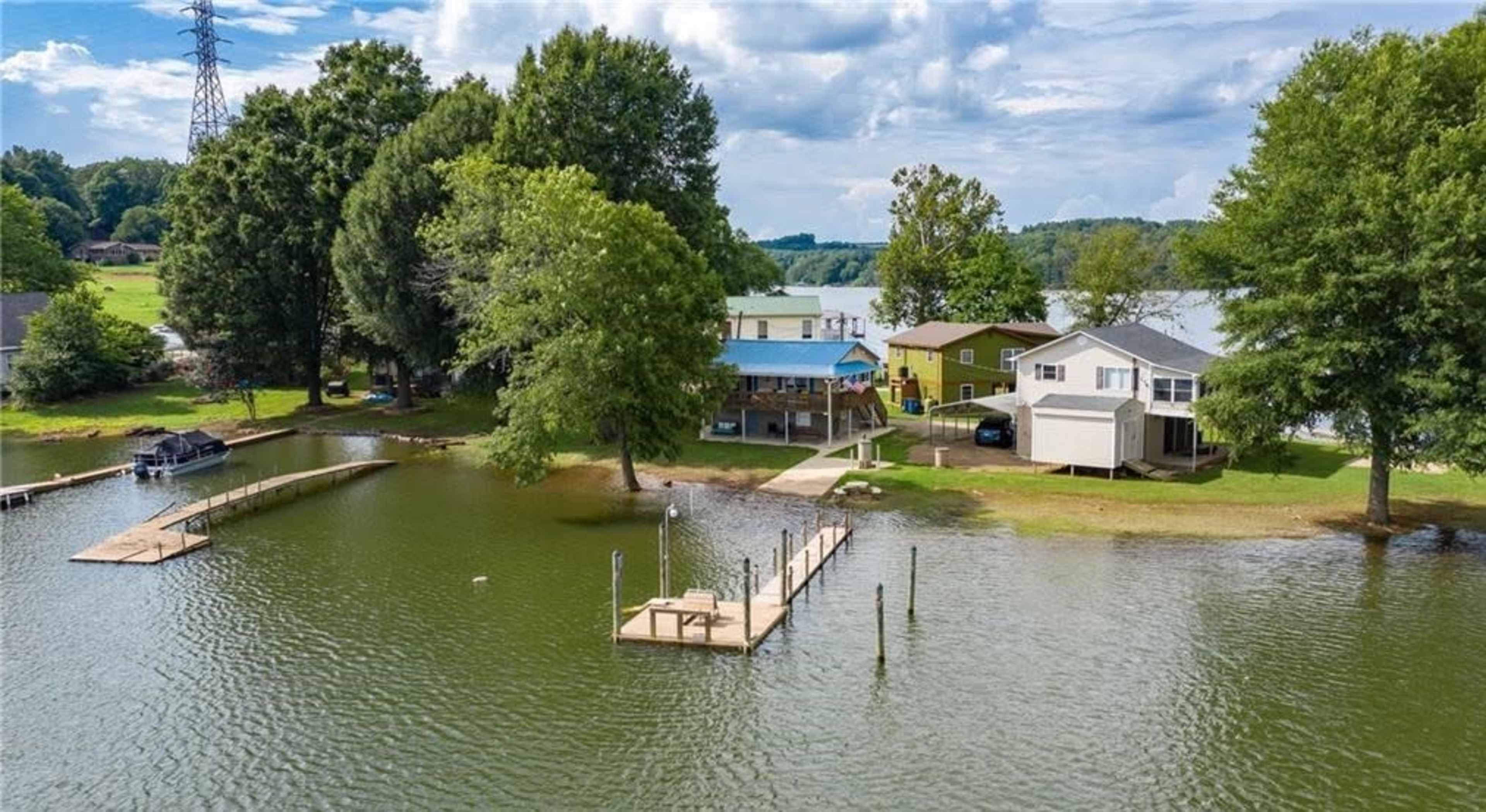 A view of a lakeside property featuring multiple buildings, docks, and lush trees under a partly cloudy sky.