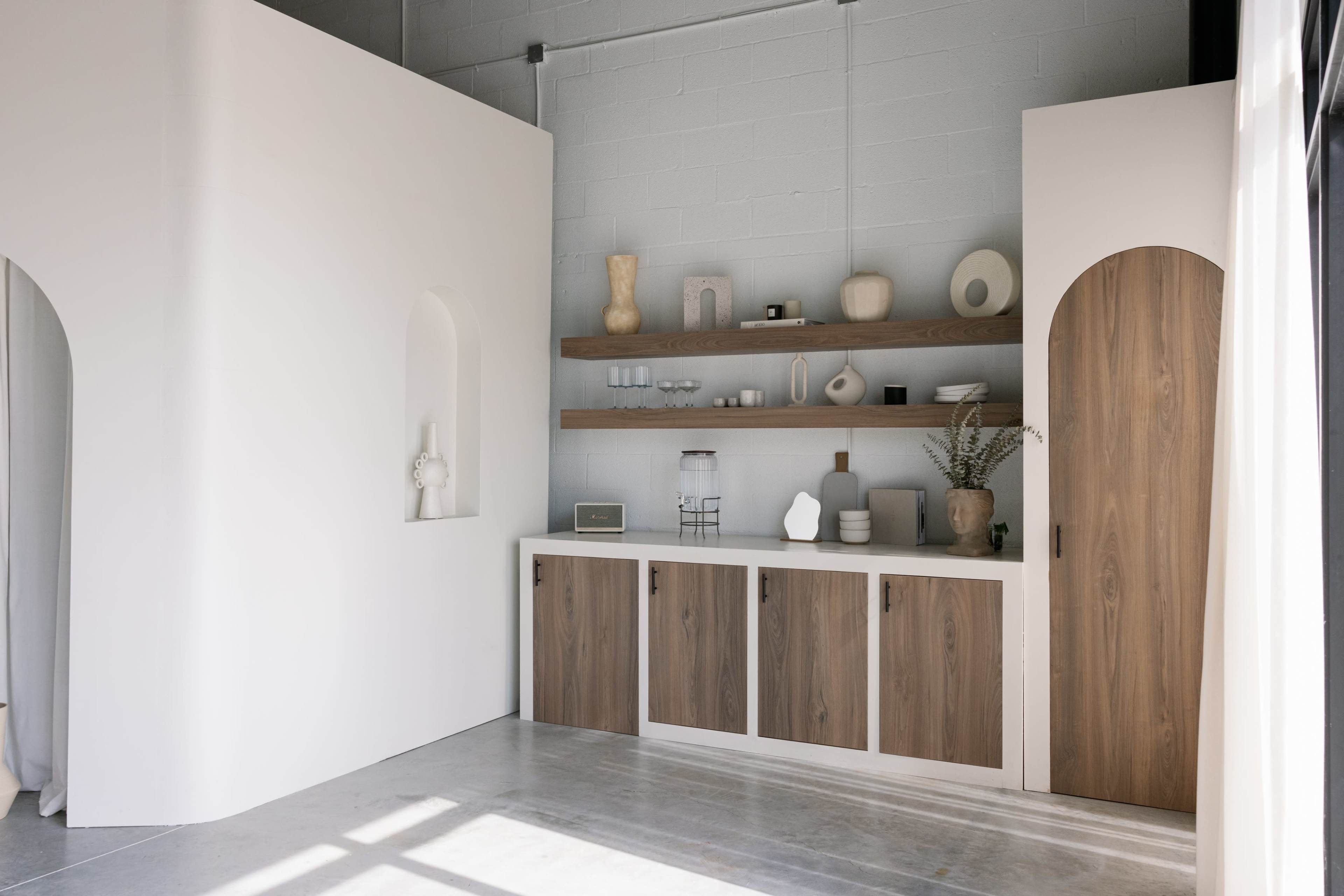 A minimalist interior space featuring a white wall with an arched niche, a wooden shelf displaying decorative items, and a storage cabinet with wooden doors.