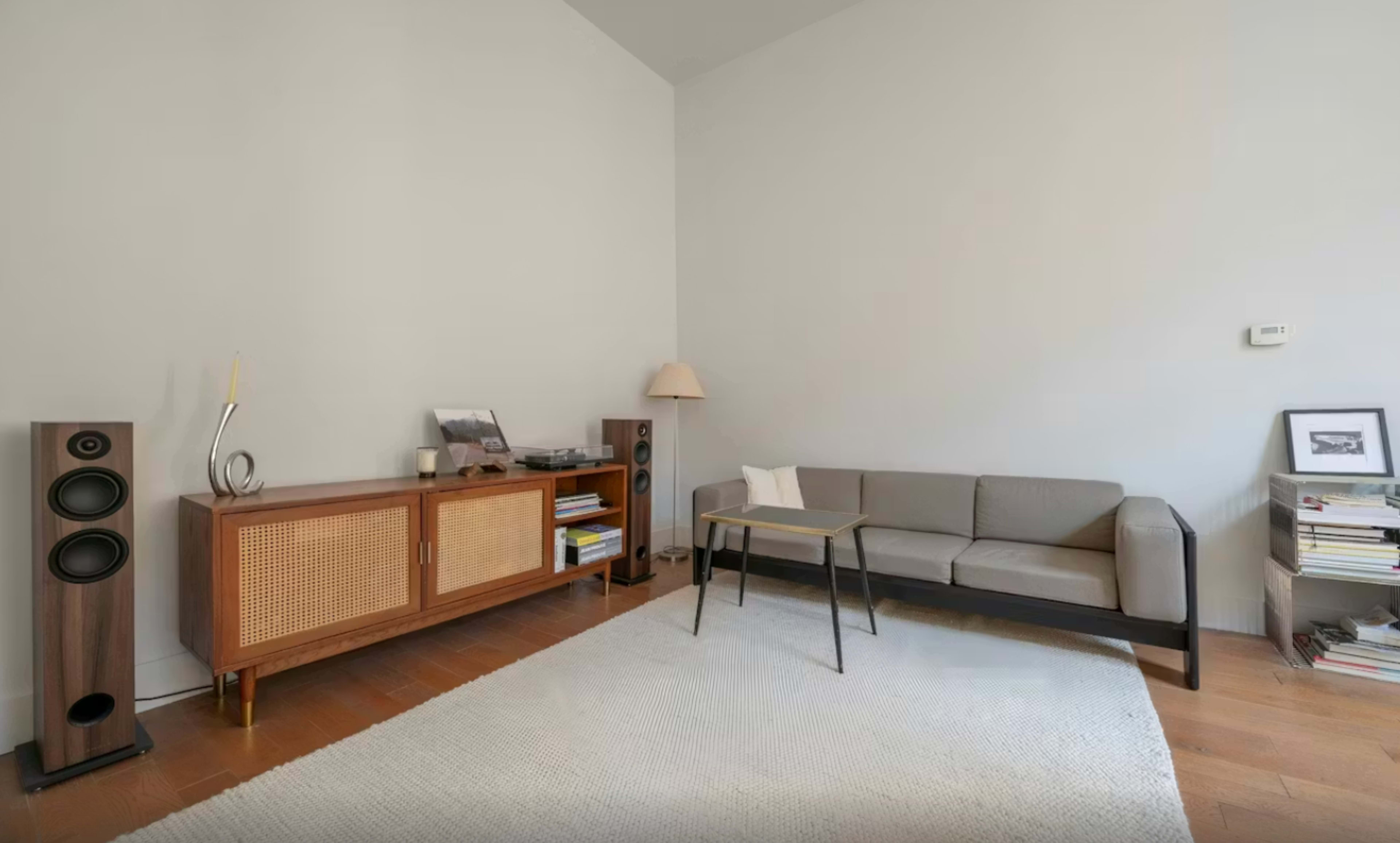 The image features a minimalist living room with a gray sofa, a wooden cabinet, a small lamp, and bookshelves against light-colored walls.
