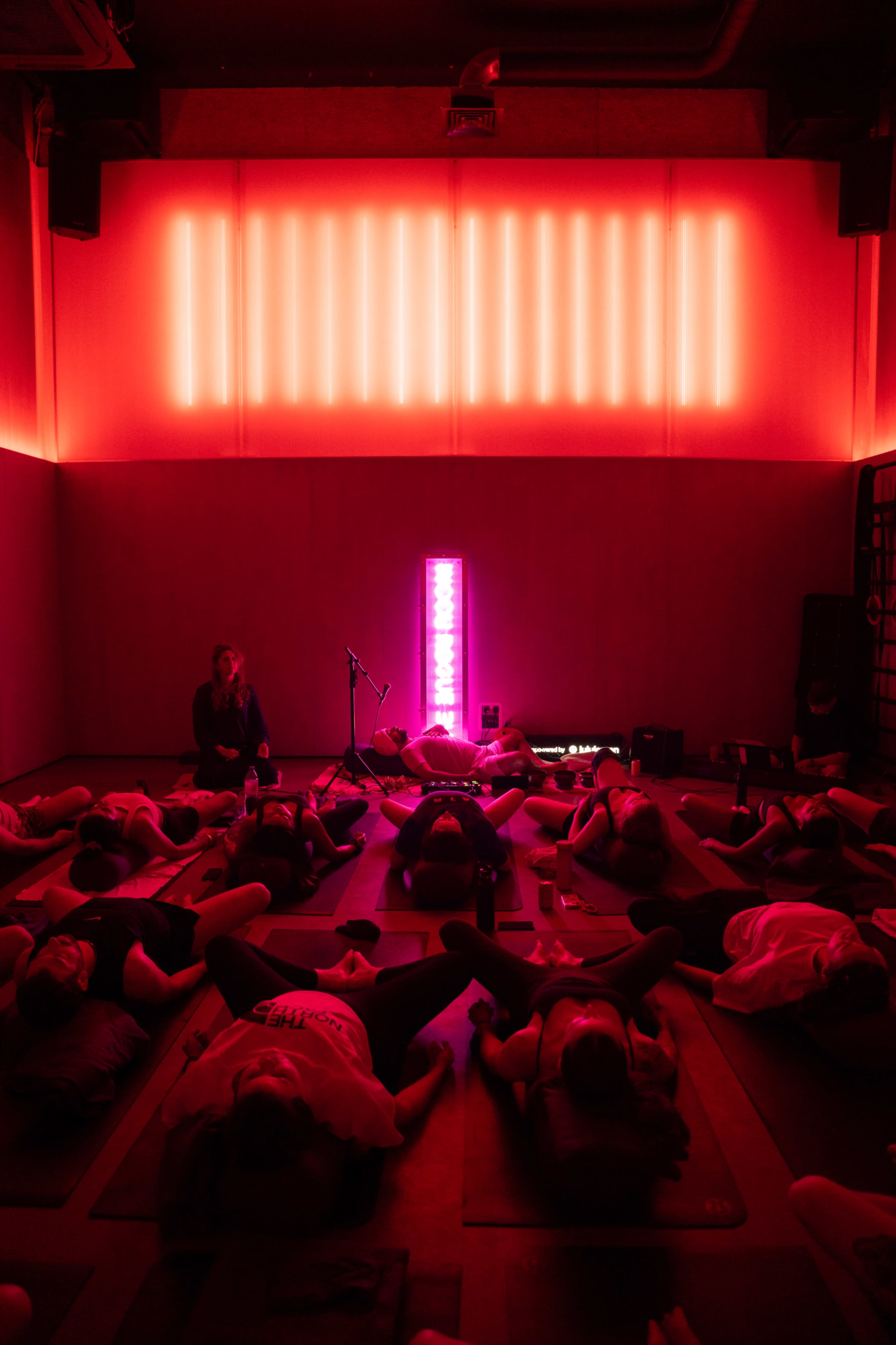 A group of individuals lies on yoga mats in a dimly lit room with red lighting, while an instructor stands in front of them.