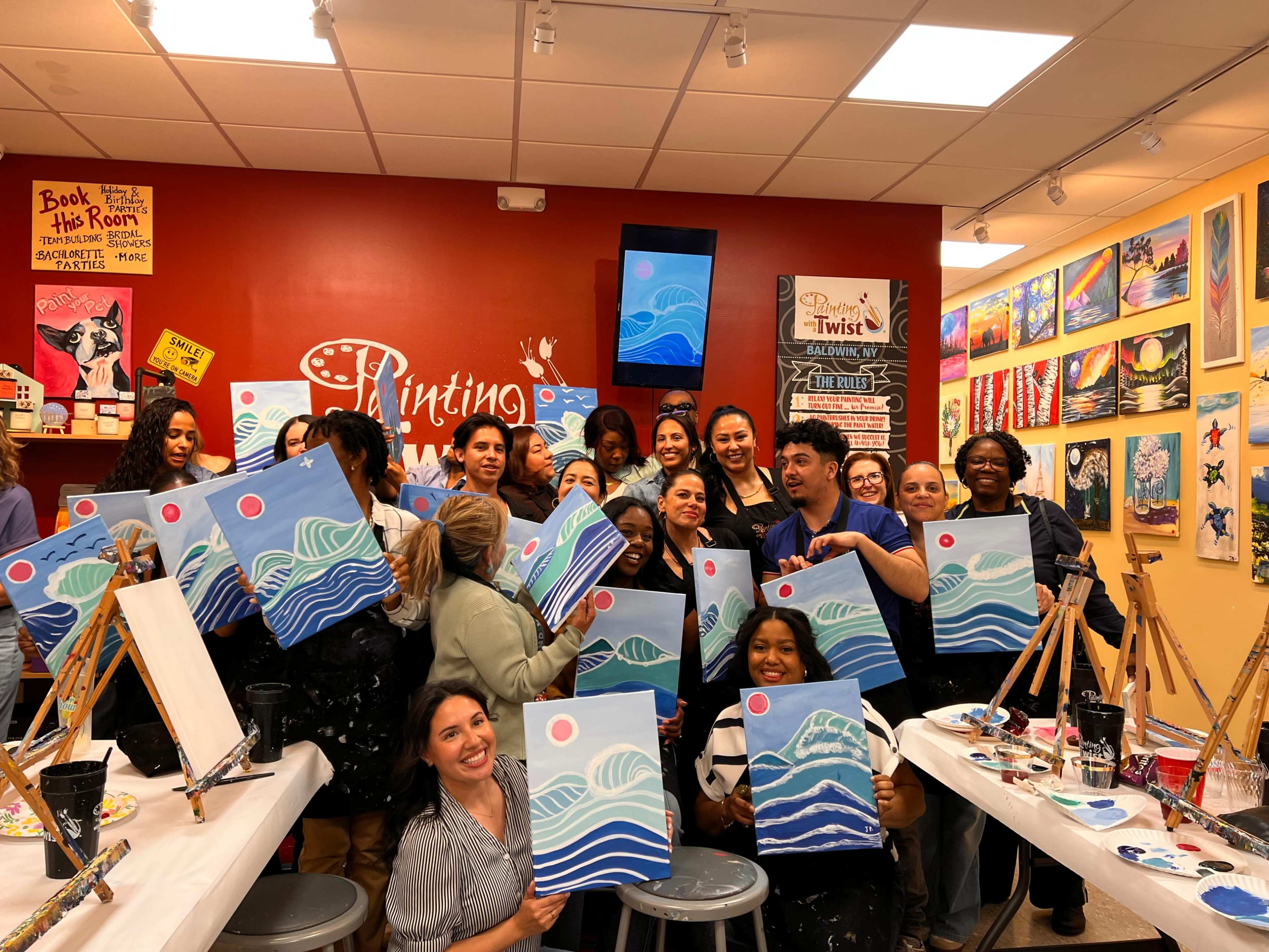 A large group of people stands in a brightly lit painting studio, each holding up their completed canvas artwork featuring waves and a red sun.