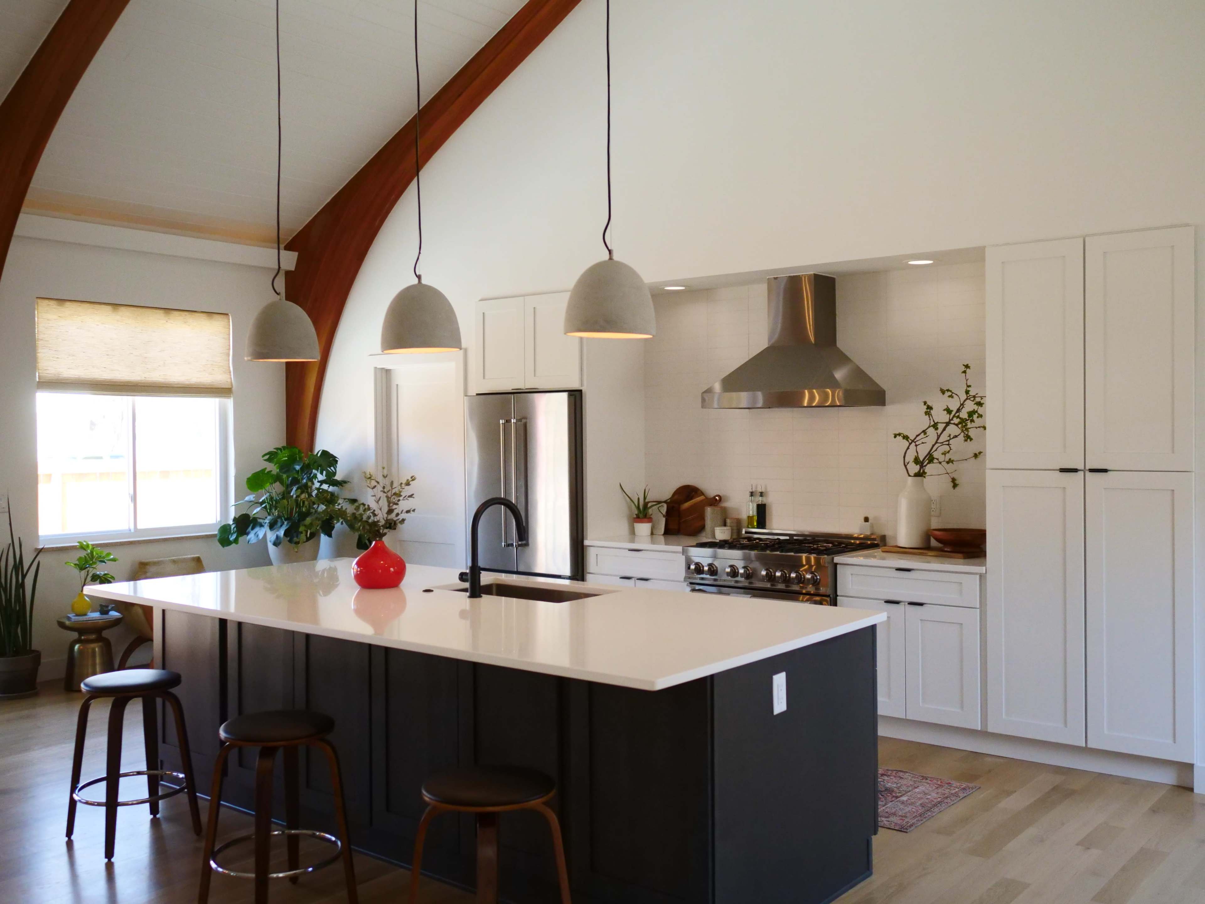 A modern kitchen featuring a large island with bar stools, stainless steel appliances, and pendant lights hanging from a high ceiling with wooden beams.