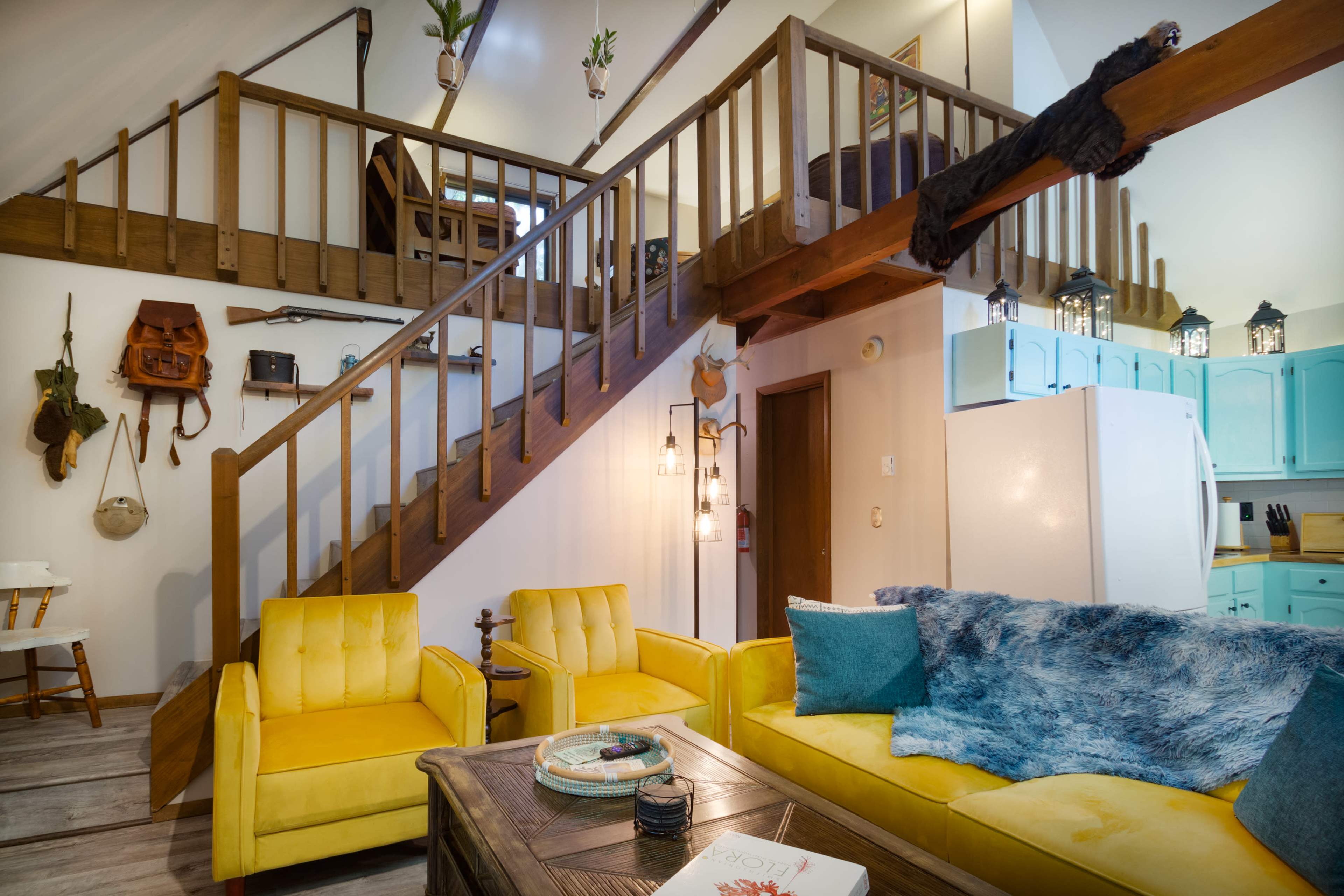 The image shows a cozy interior with a wooden staircase leading to an upper level, yellow couches, and a kitchen area featuring light blue cabinets.