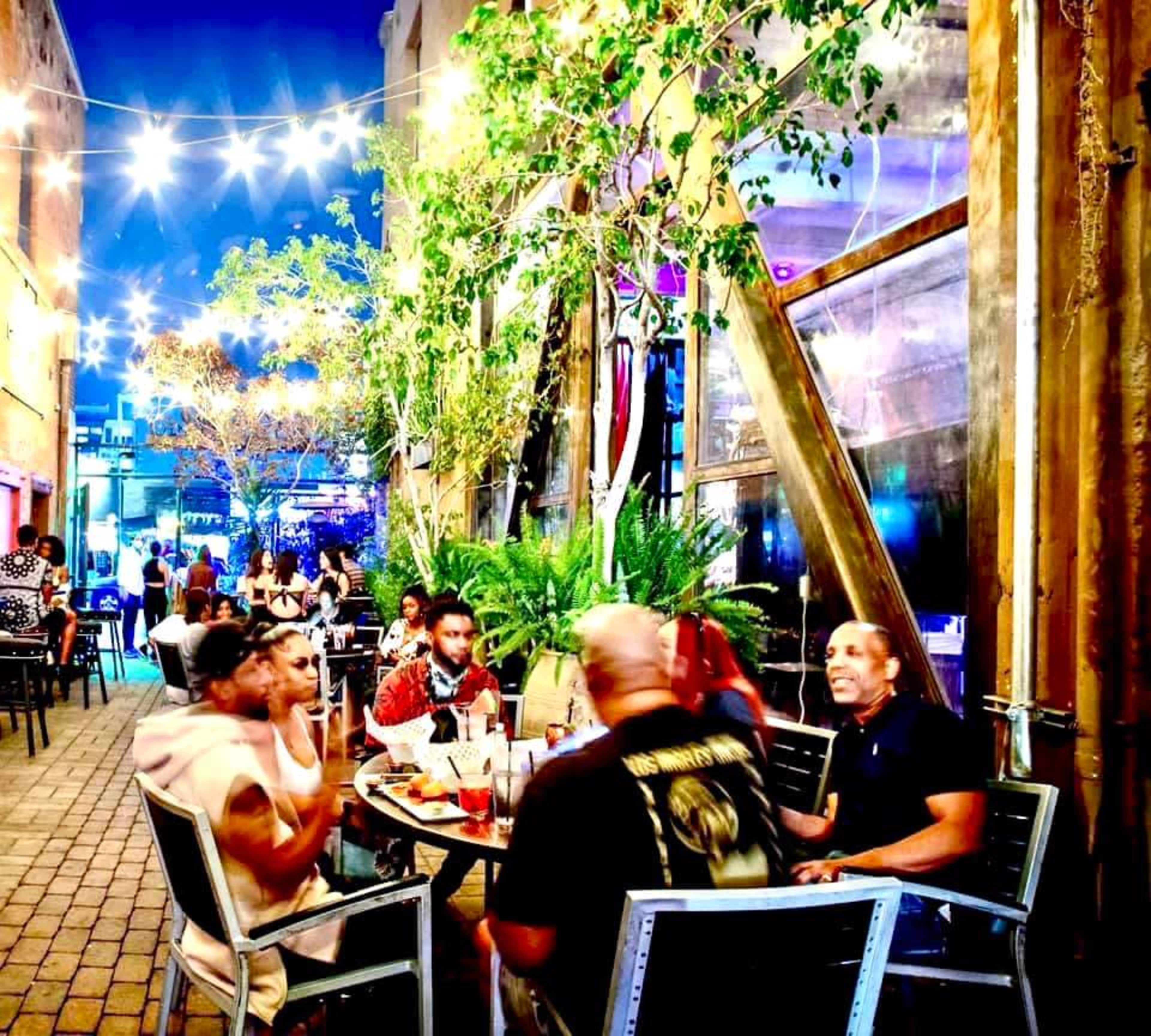 A group of people sit at tables in a well-lit outdoor restaurant area surrounded by greenery and string lights.