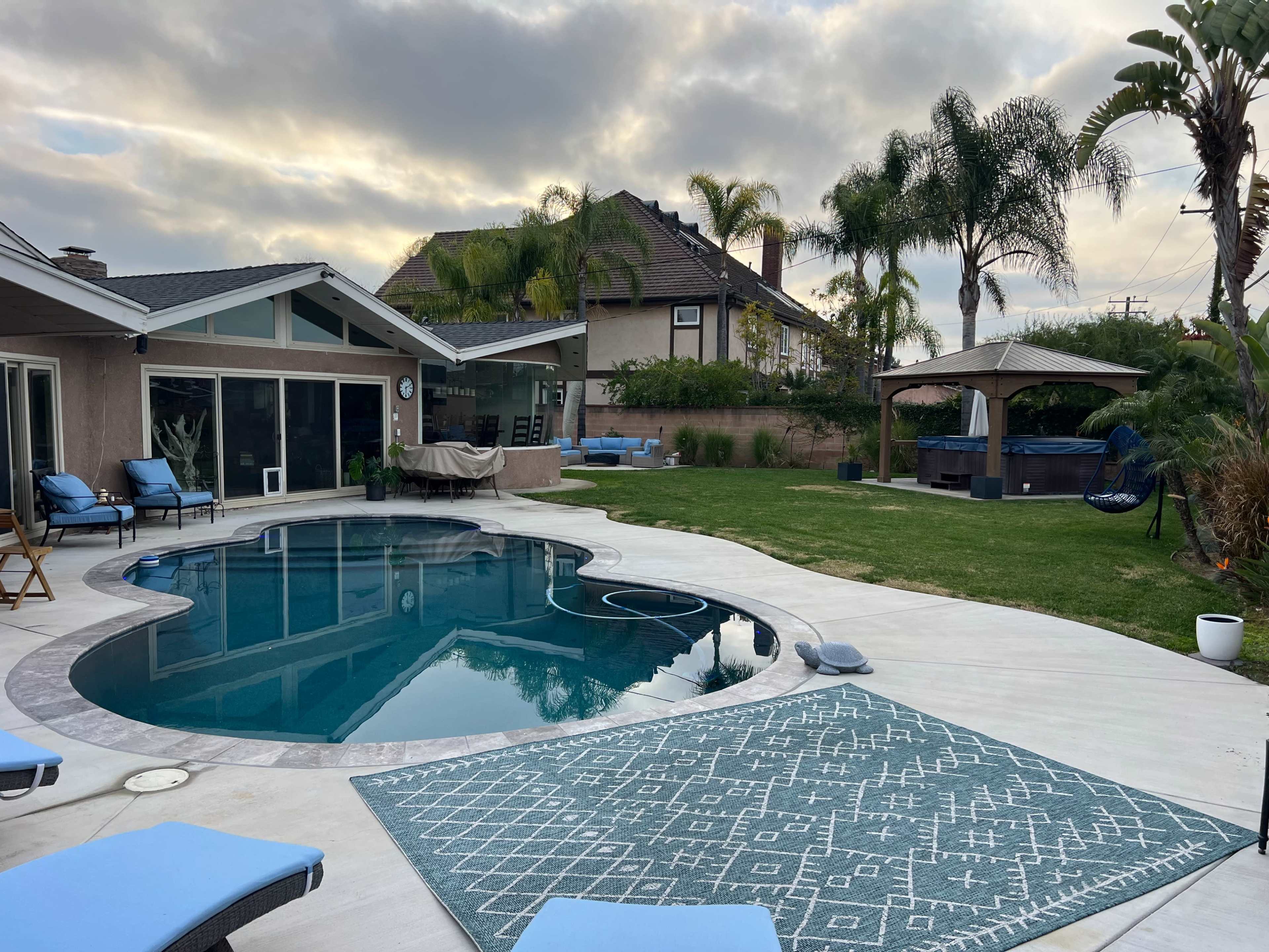 A backyard with a swimming pool, lounge chairs, and a well-maintained lawn surrounded by palm trees and a house.