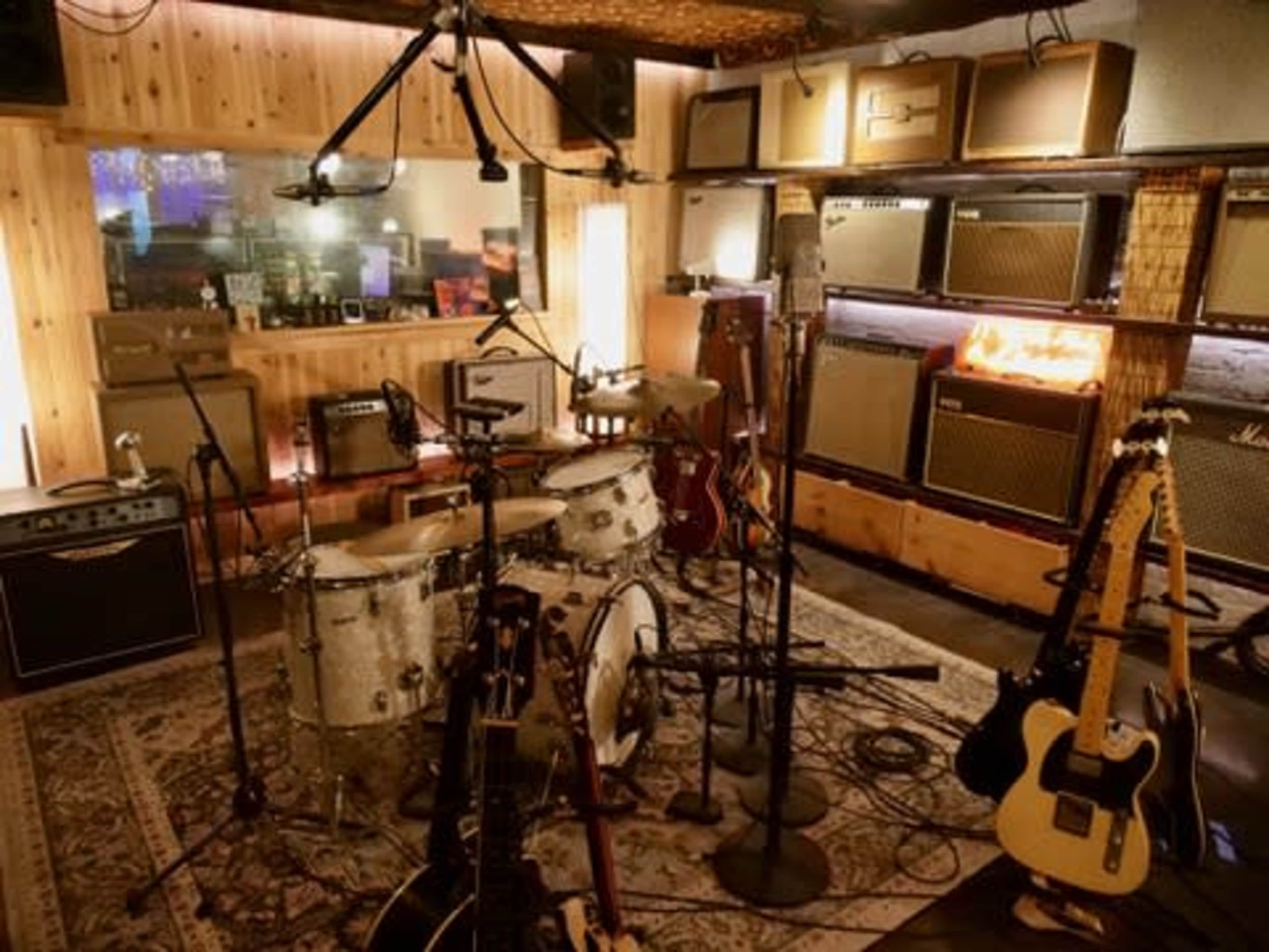 The image shows a recording studio filled with various musical instruments, including a drum set and multiple guitar amplifiers on display.
