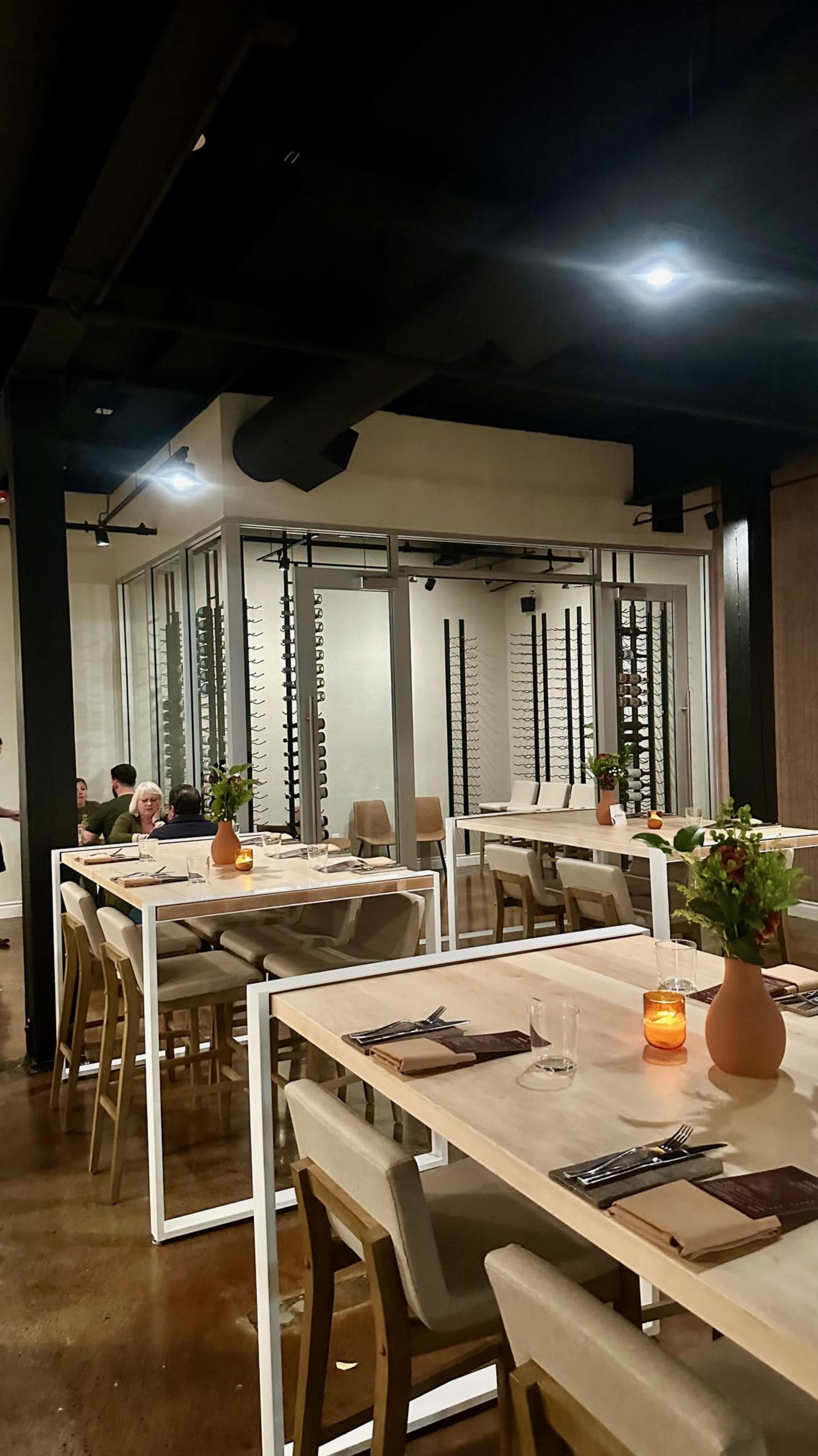 The image shows a modern restaurant interior with minimalistic wooden tables, light-colored chairs, and a prominent wine storage area visible through glass walls.