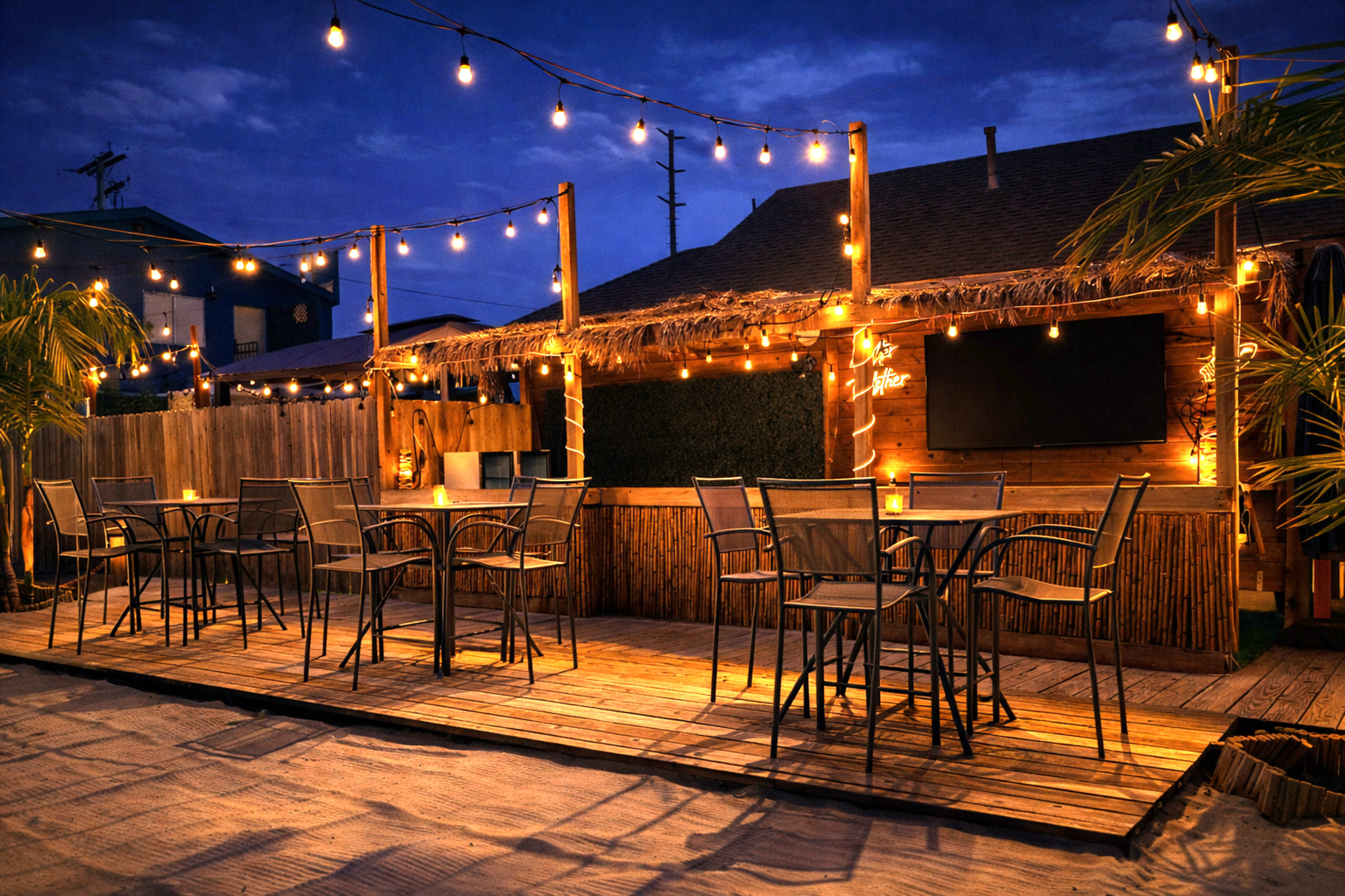 The image shows an outdoor seating area with tables and chairs, surrounded by string lights and a thatched-roof structure, set against a twilight sky.
