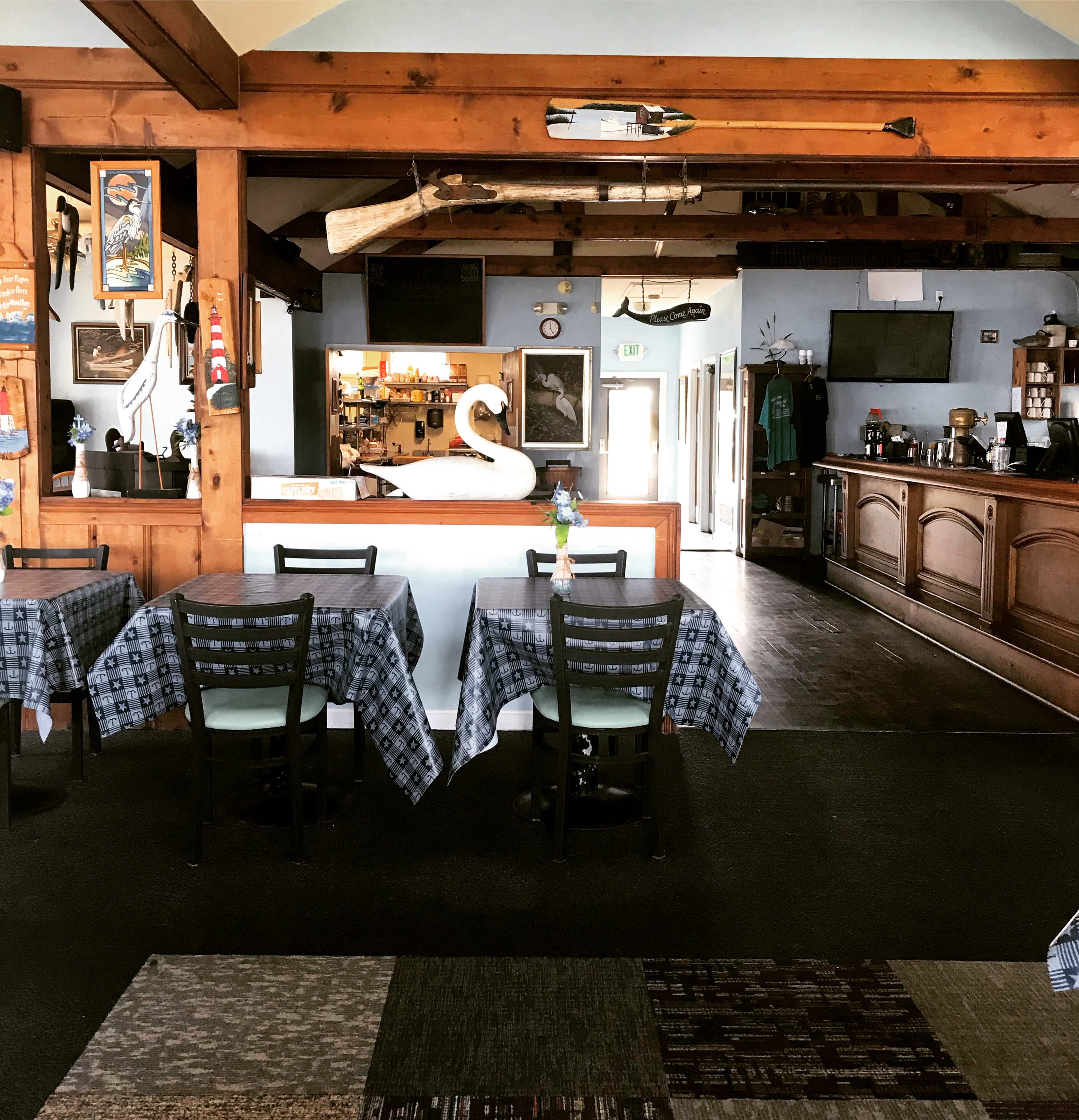 An empty restaurant interior with wooden beams, tables covered with checkered tablecloths, and a small bar area in the background.