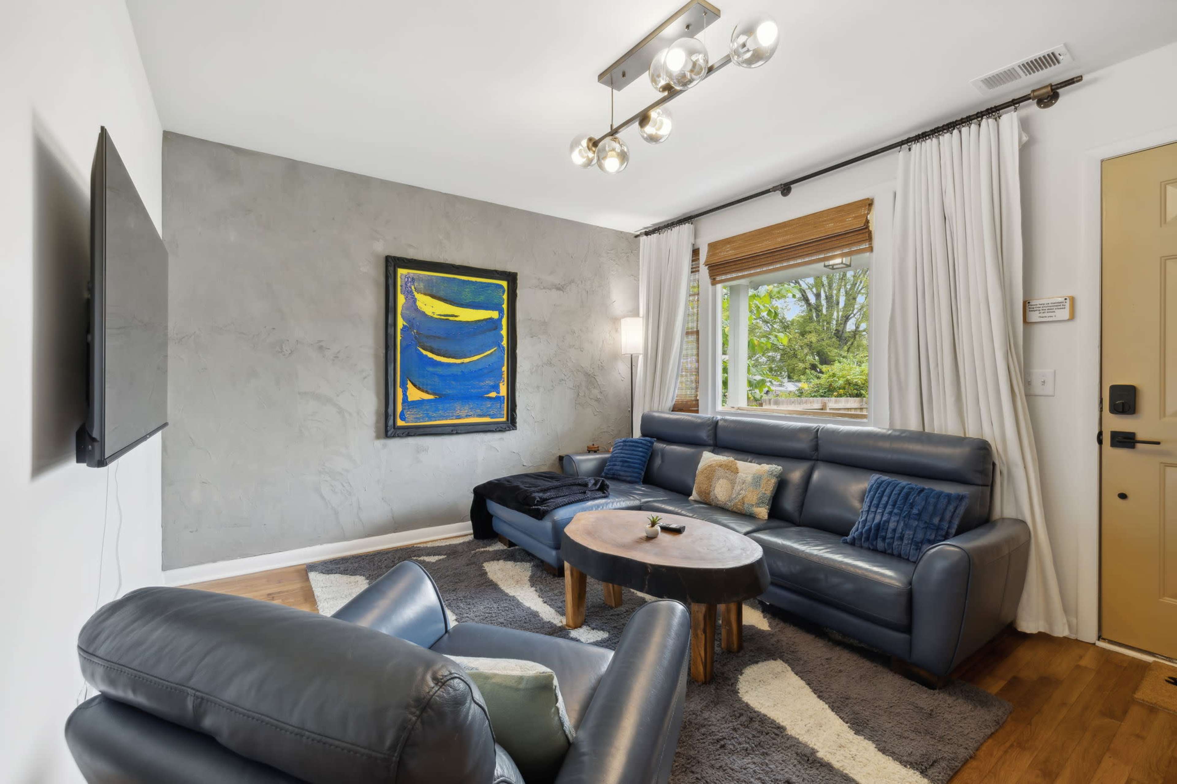 The image shows a cozy living room with a gray accent wall, a large blue painting, and a dark leather sectional sofa facing a wall-mounted TV.