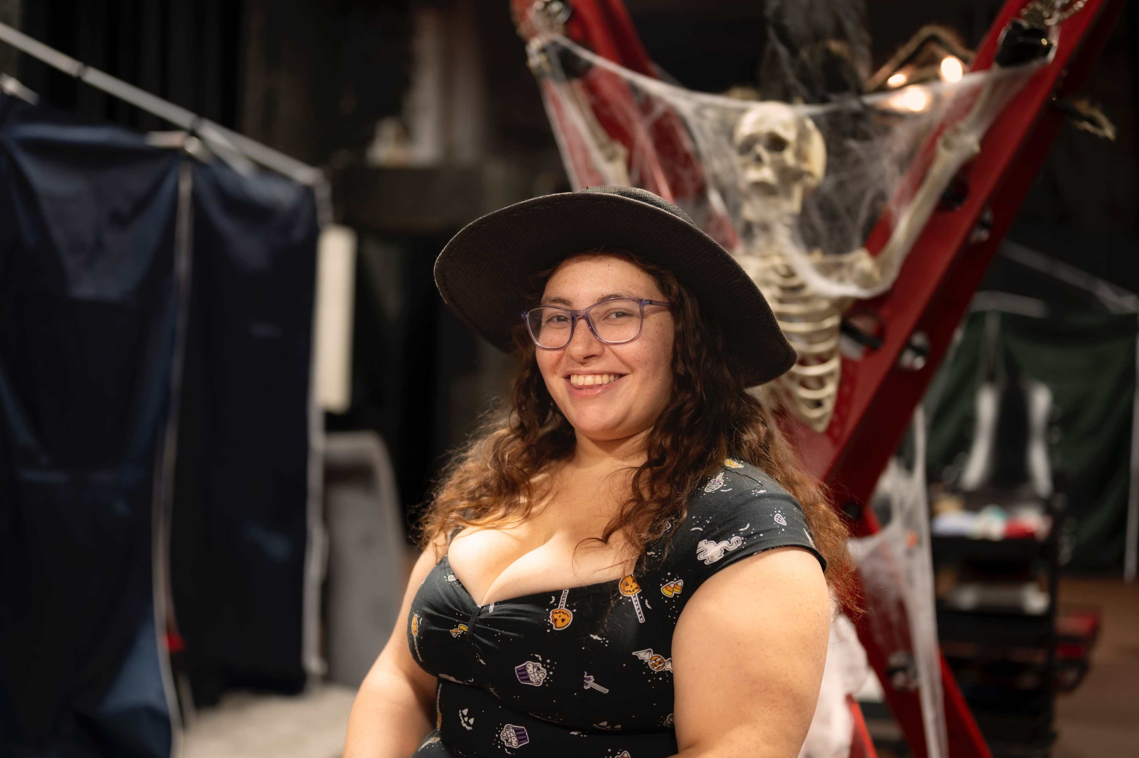 A woman wearing a black hat and a patterned dress poses in front of a skeleton decorated with cobwebs.