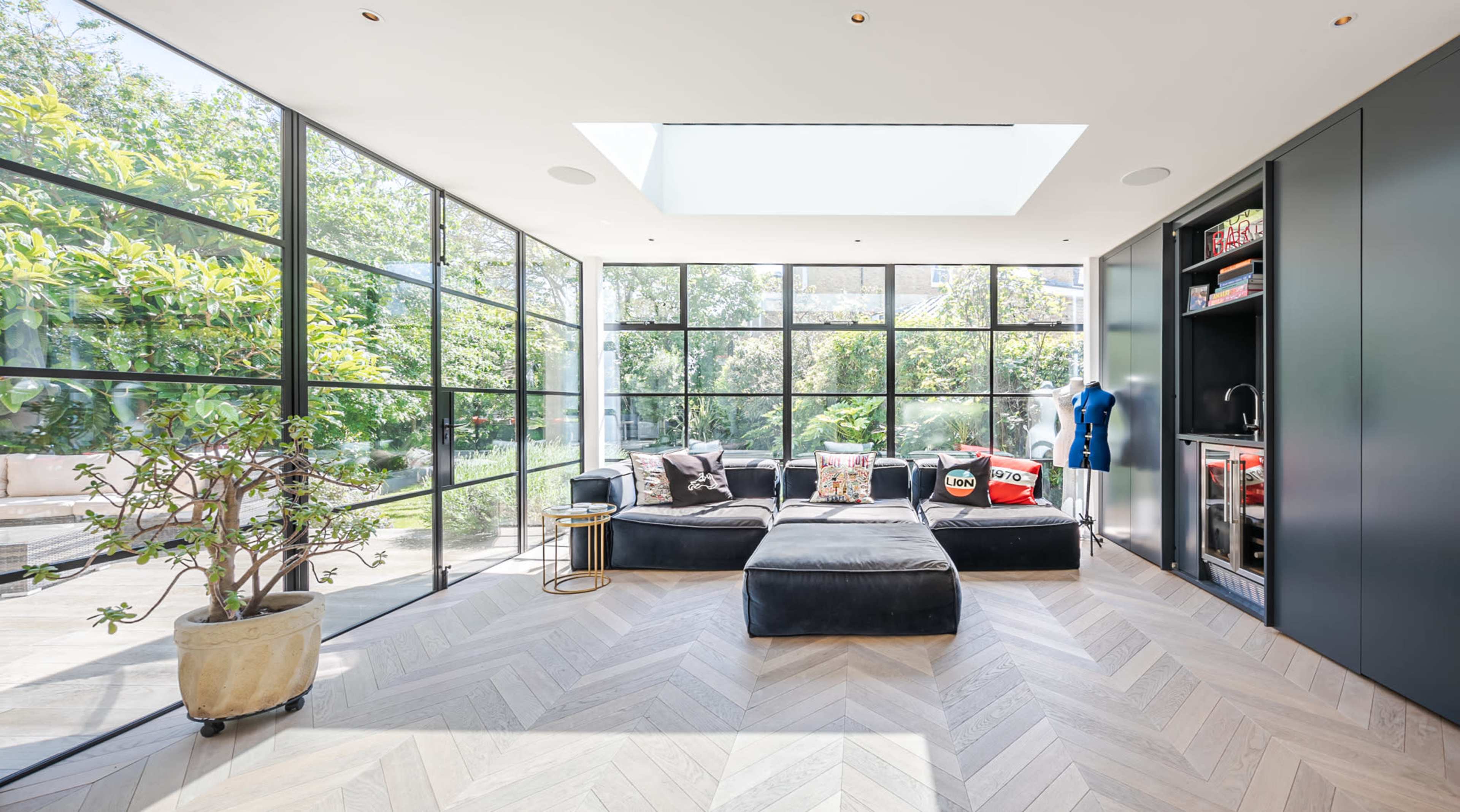 A modern living room features large windows that offer a view of greenery, with a black sectional couch and a small side table.