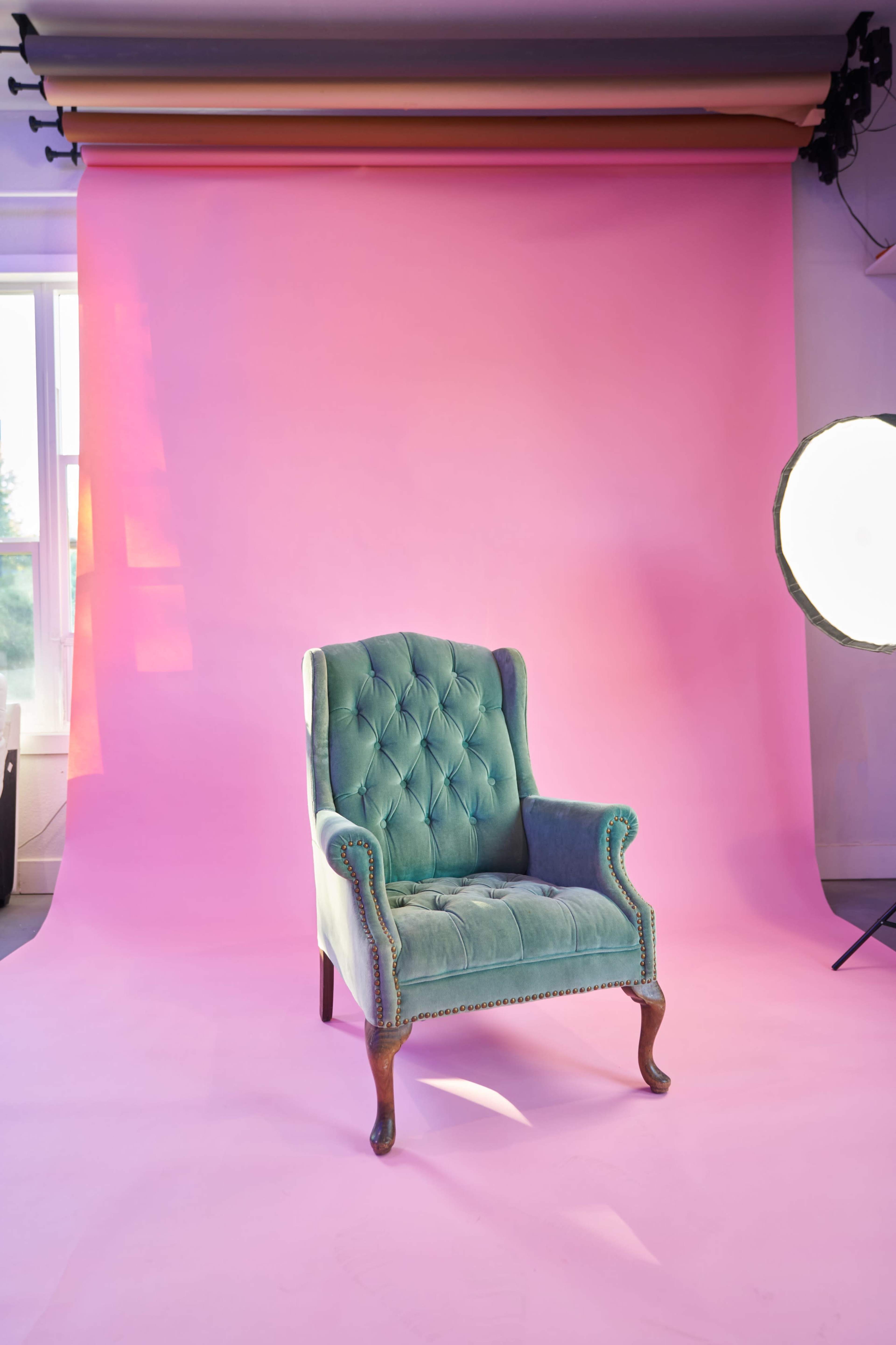 A vintage blue velvet armchair is positioned in front of a bright pink backdrop in a well-lit room.