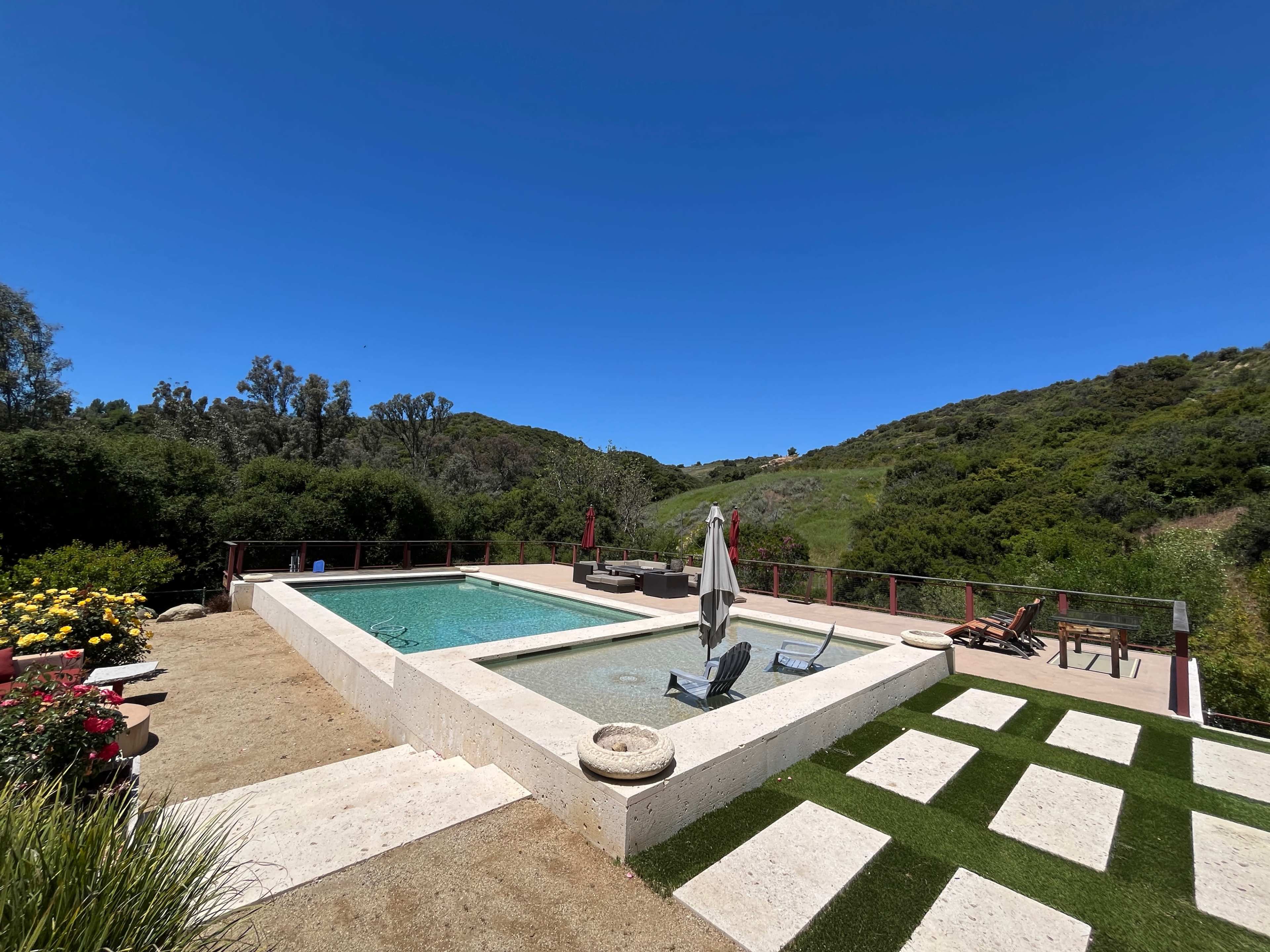The image shows a landscaped outdoor area featuring two swimming pools surrounded by greenery and a hillside.