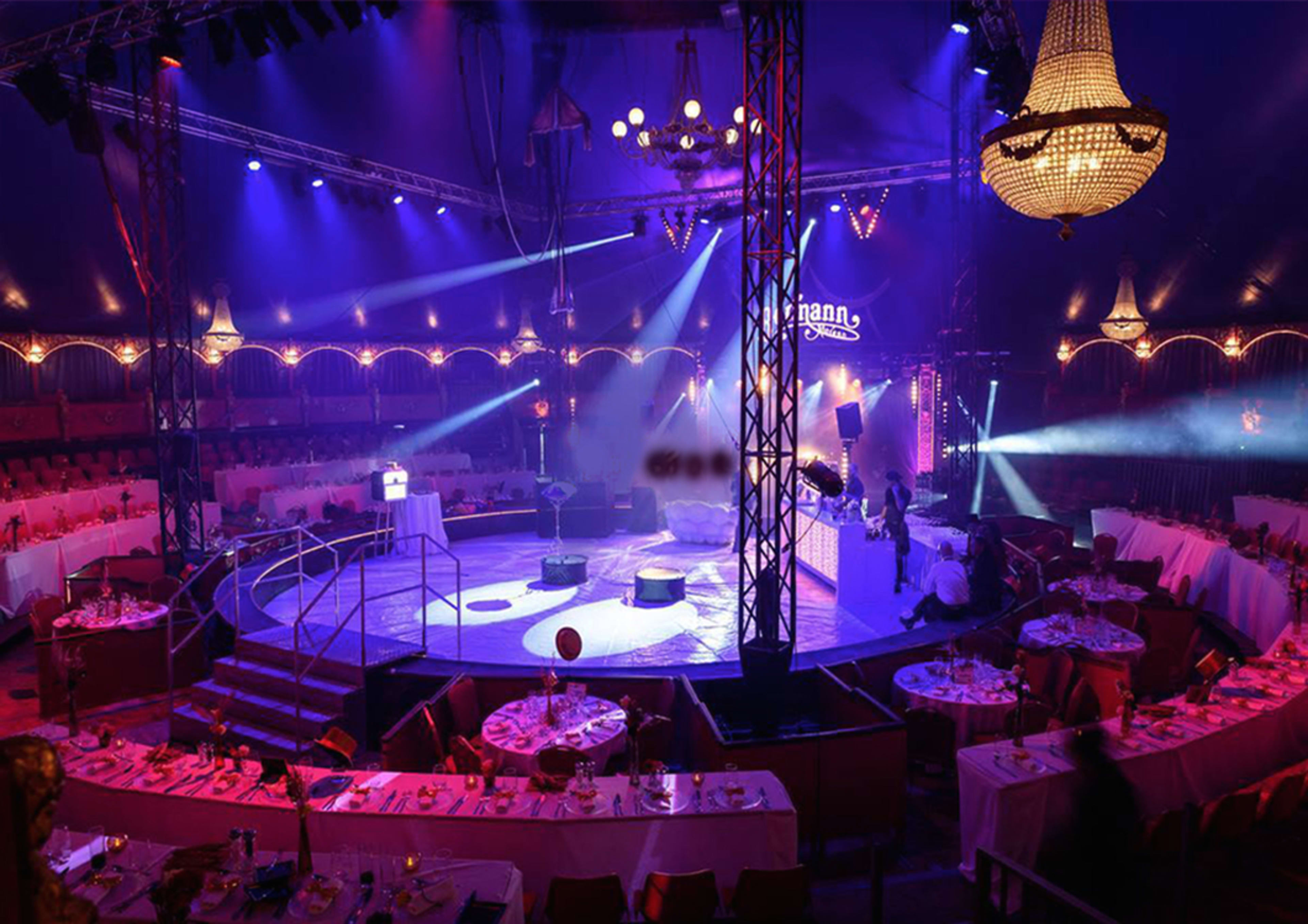 An empty circus tent with a circular performance area surrounded by tables and chairs, illuminated by stage lights.