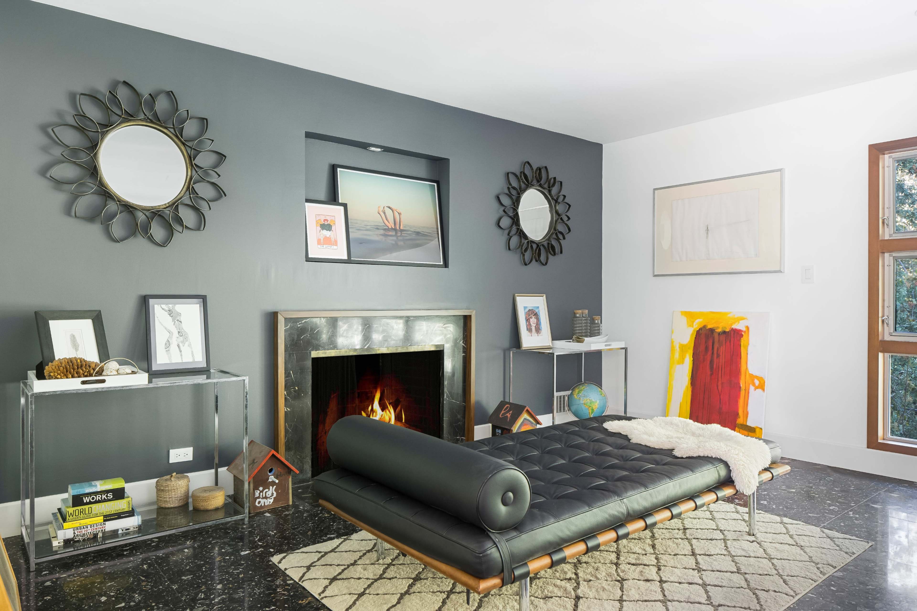 The room features a modern lounge area with a black leather daybed, a circular mirror, and a fireplace with a fire, surrounded by various art pieces and decor.