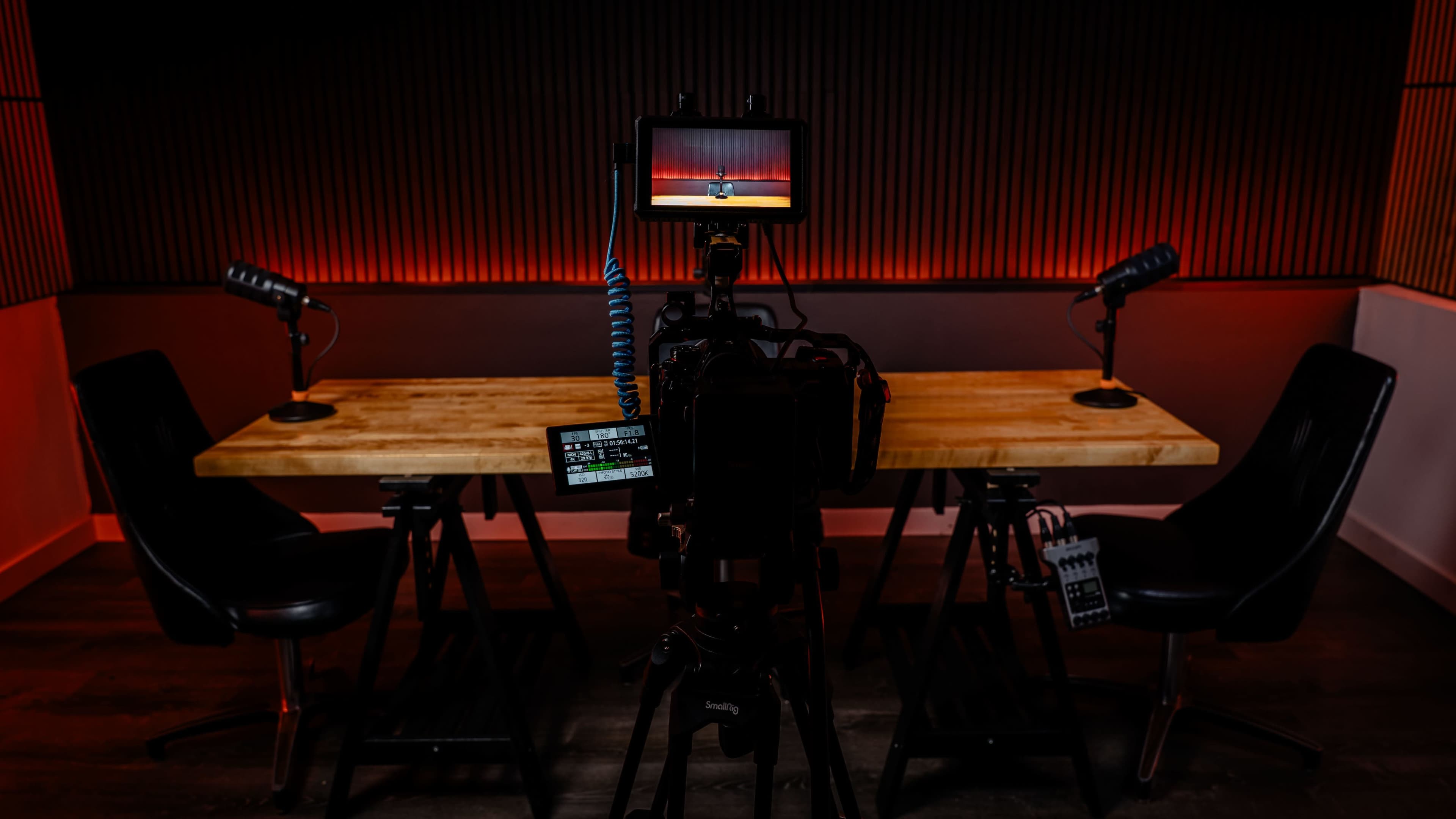 A camera is positioned in front of a wooden table set in a dimly lit studio, with microphones on either side and a screen displaying sunset imagery.