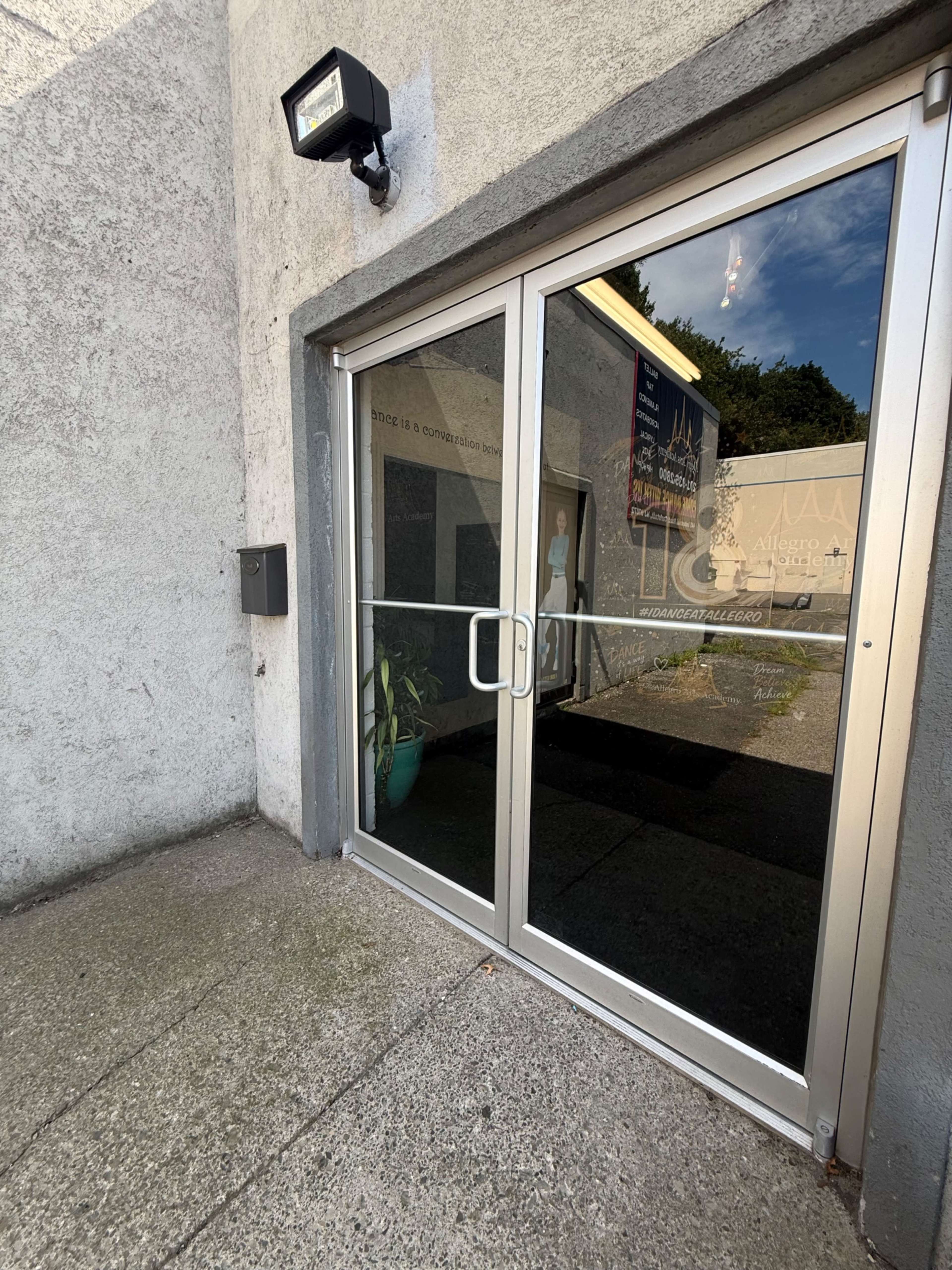 The image shows a glass double door entrance to a building, flanked by a light fixture and a mail slot on the wall beside it.
