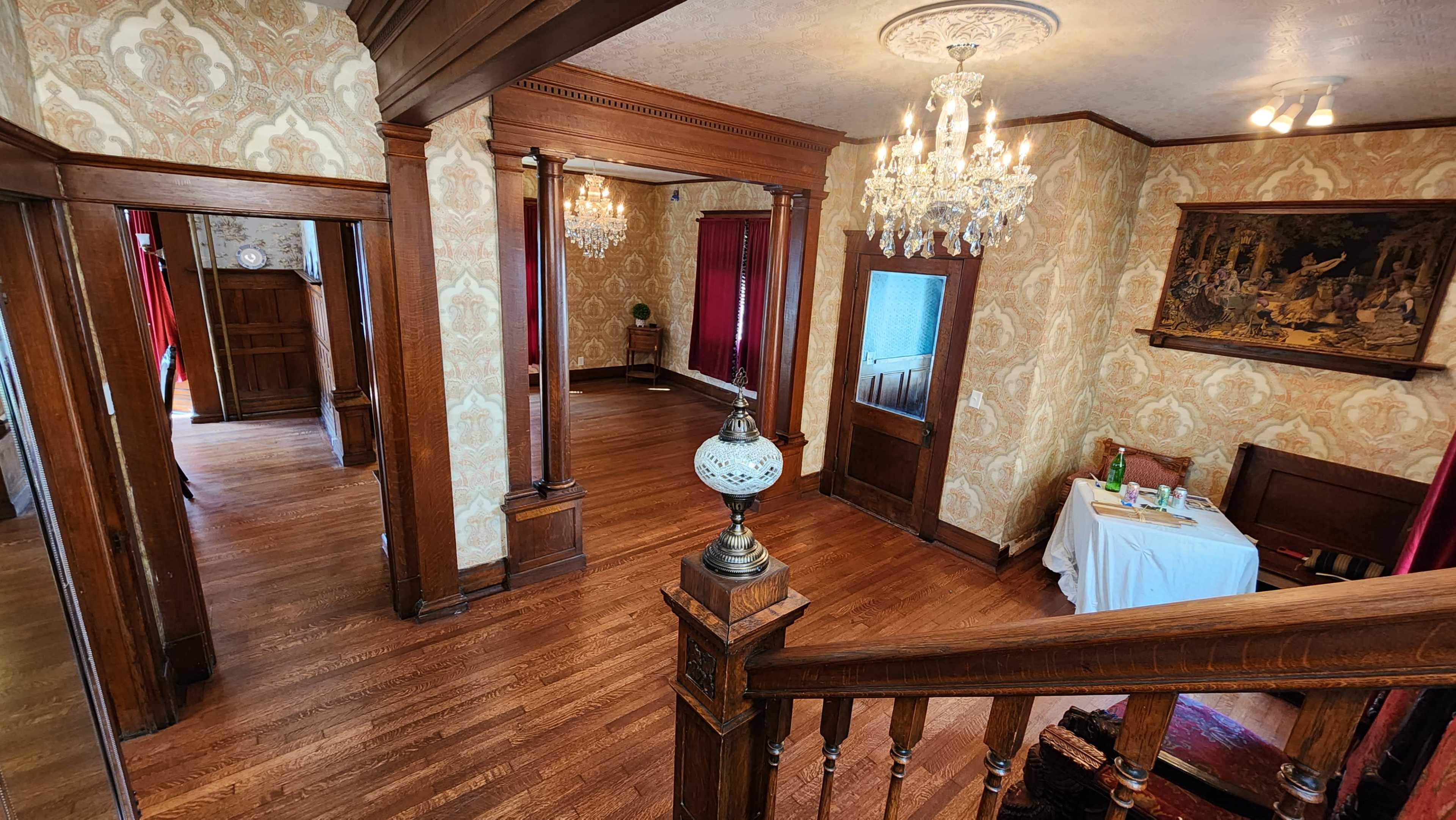 A vintage interior of a home featuring wooden pillars, chandeliers, and patterned wallpaper, with a view of a dining table in one corner.