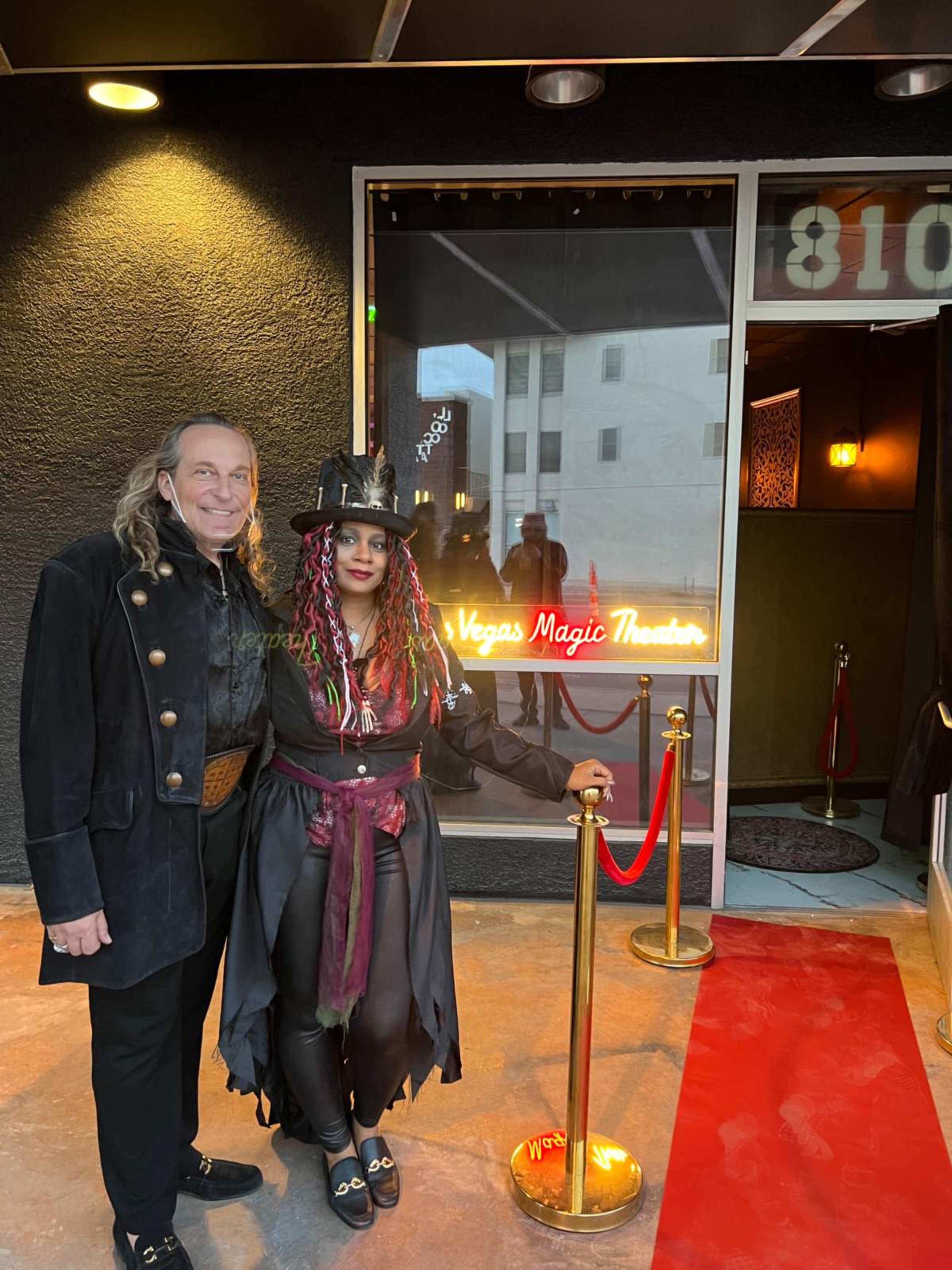 A man and a woman stand in front of the entrance to a magic theater, with a red carpet and a sign that reads "Las Vegas Magic Theater."