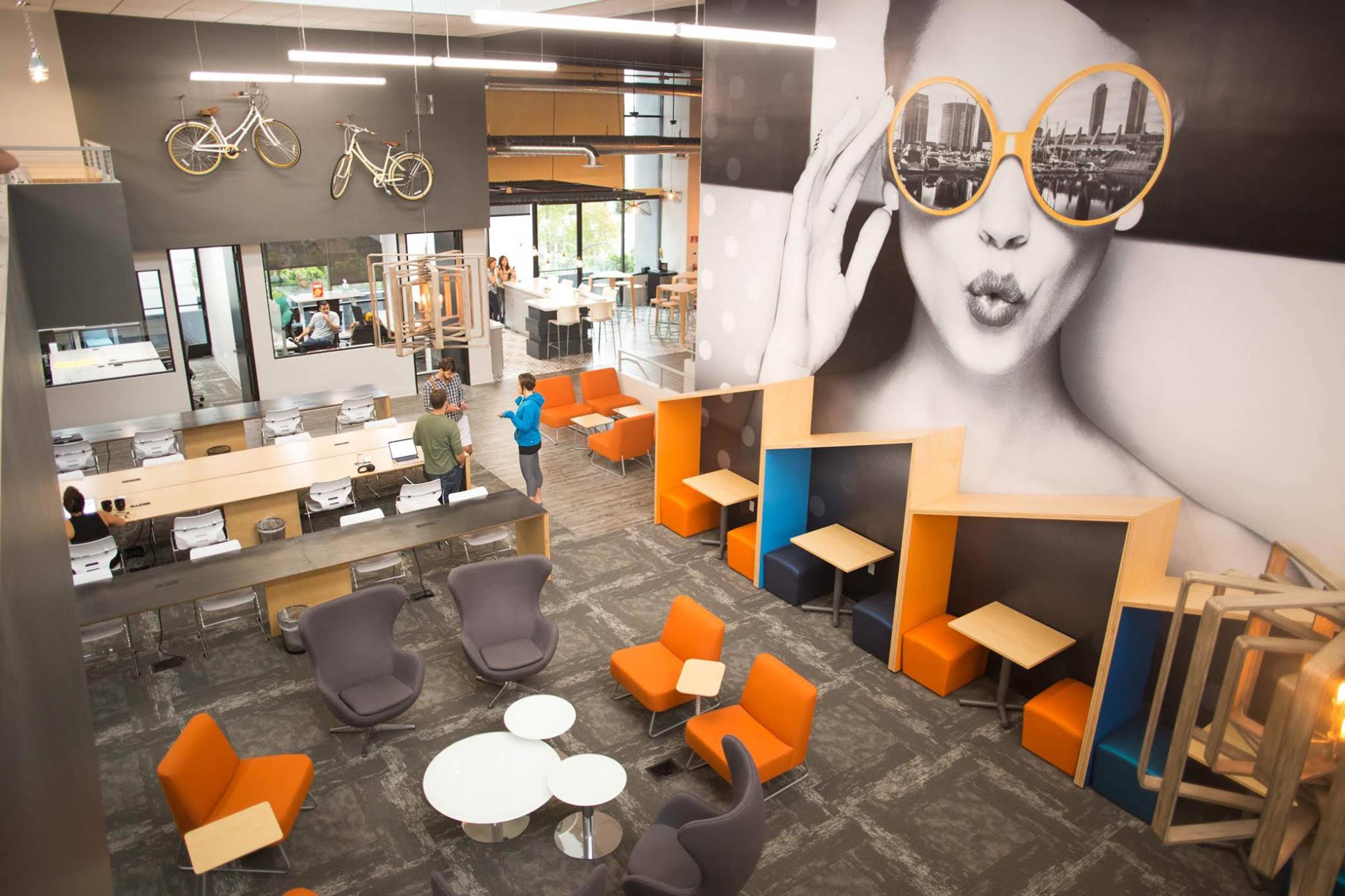 A modern, open-concept office space features a large mural of a woman with sunglasses, seating areas with orange and gray furniture, and a reception area with a minimalist design.