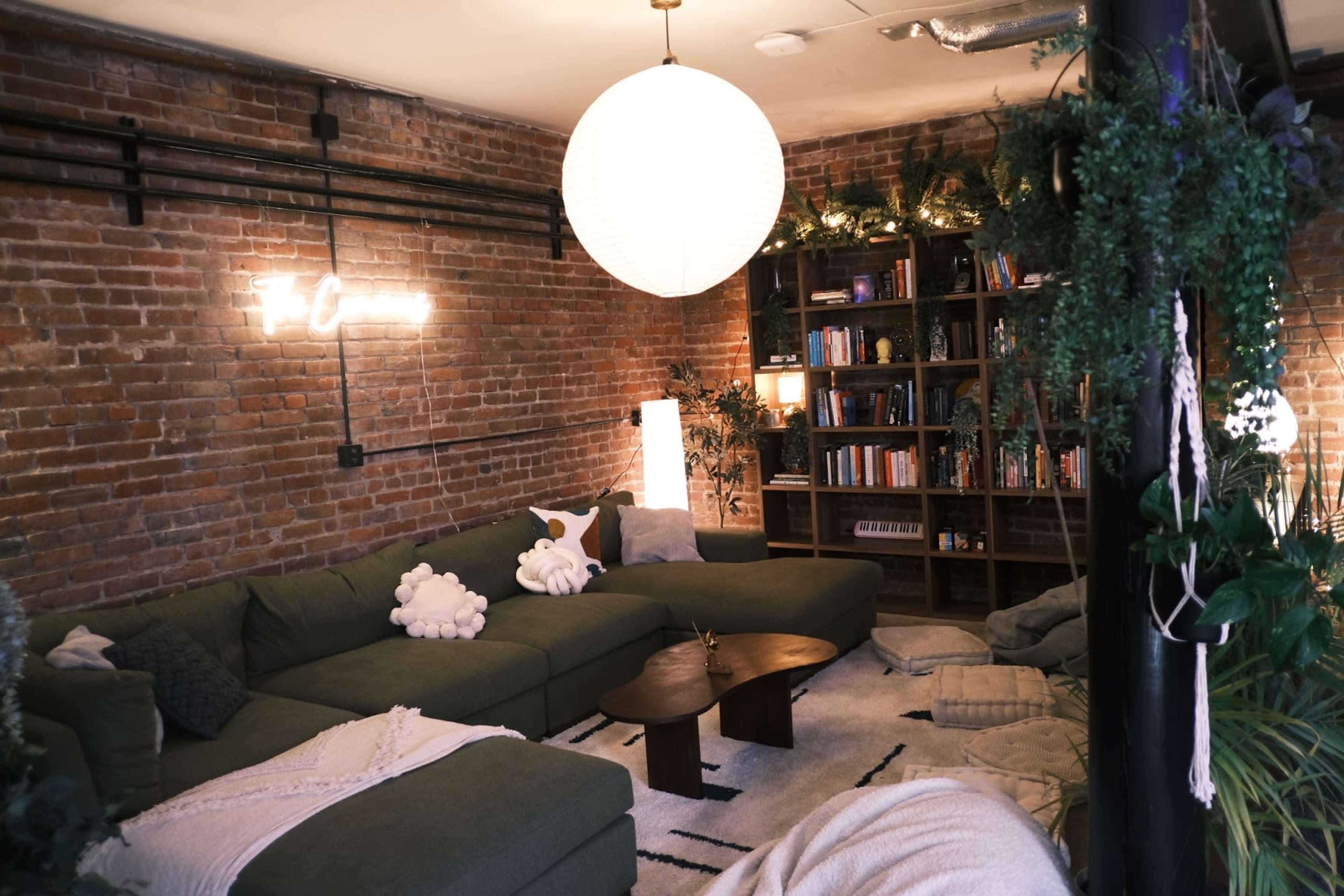 A cozy living room with a large green sectional sofa, a round coffee table, bookshelves filled with books, and plants adorning the space, all under a warm pendant light.