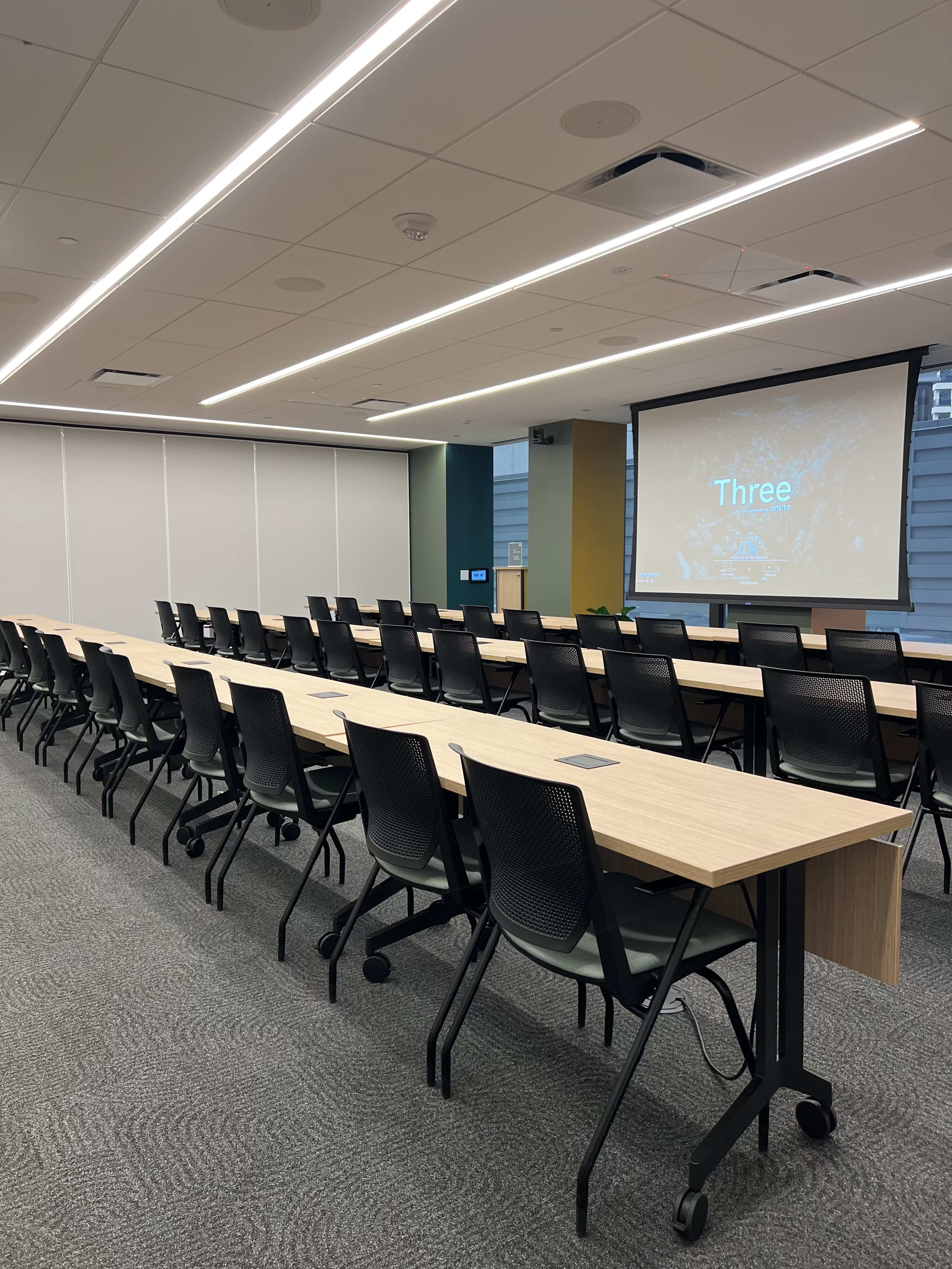 The image shows a modern conference room with multiple rows of desktop tables and office chairs, and a presentation screen displaying the word "Three."