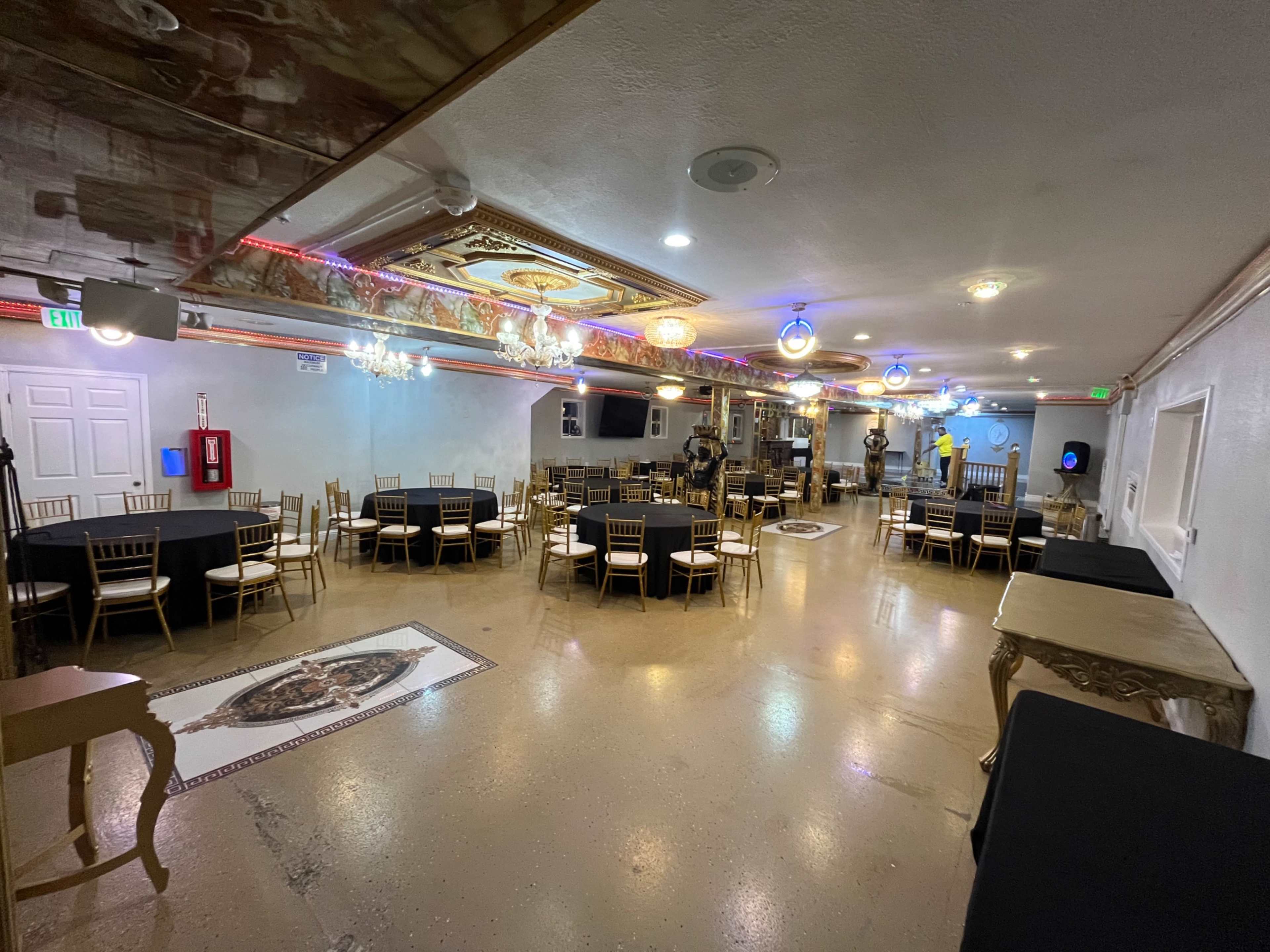 The image shows a spacious event room with black tables and gold chairs arranged neatly, illuminated by various lights and chandeliers.
