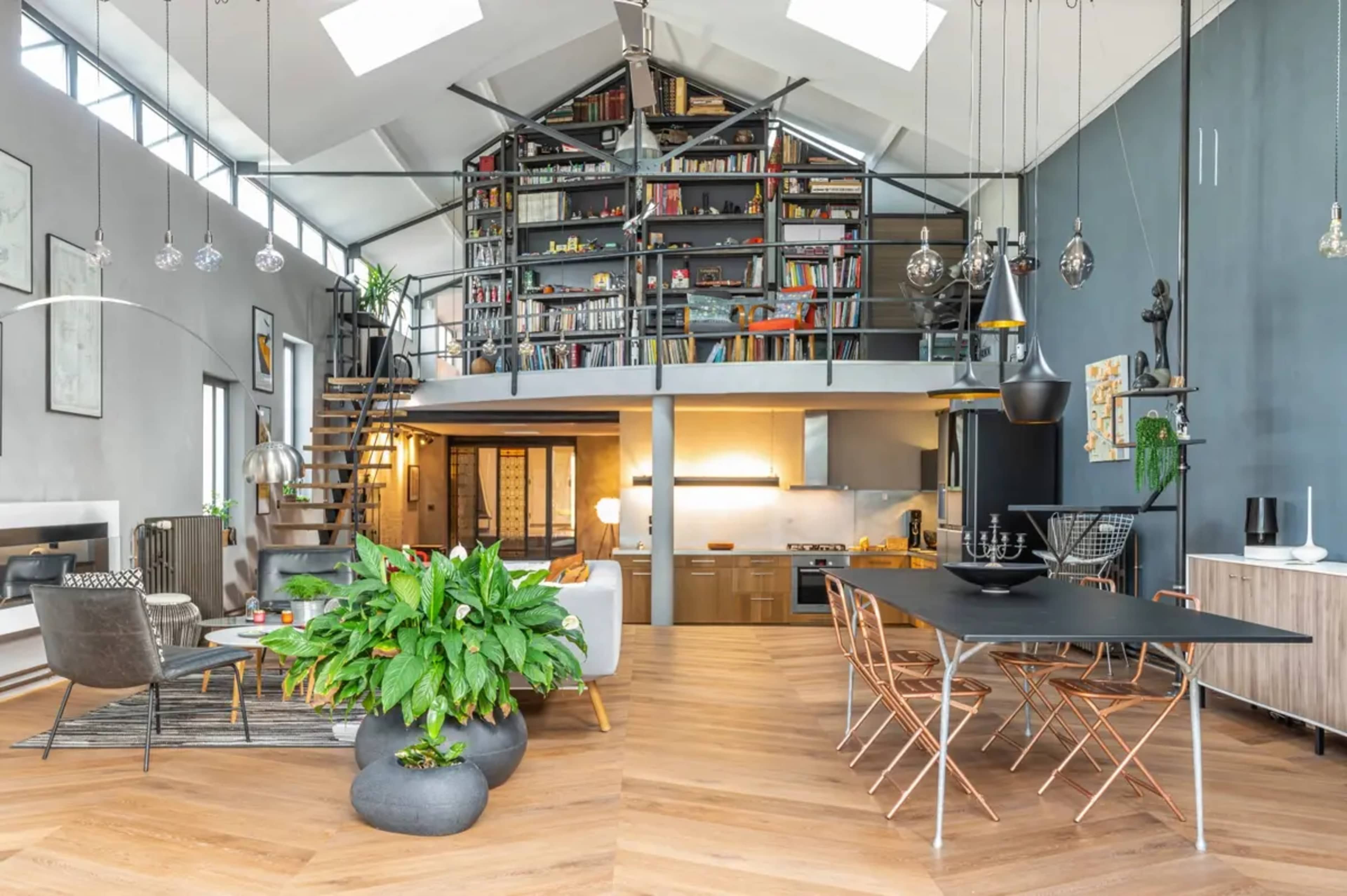 A modern open-concept living space with a dining area, kitchen, and a lofted bookshelf, featuring large windows and wooden flooring.