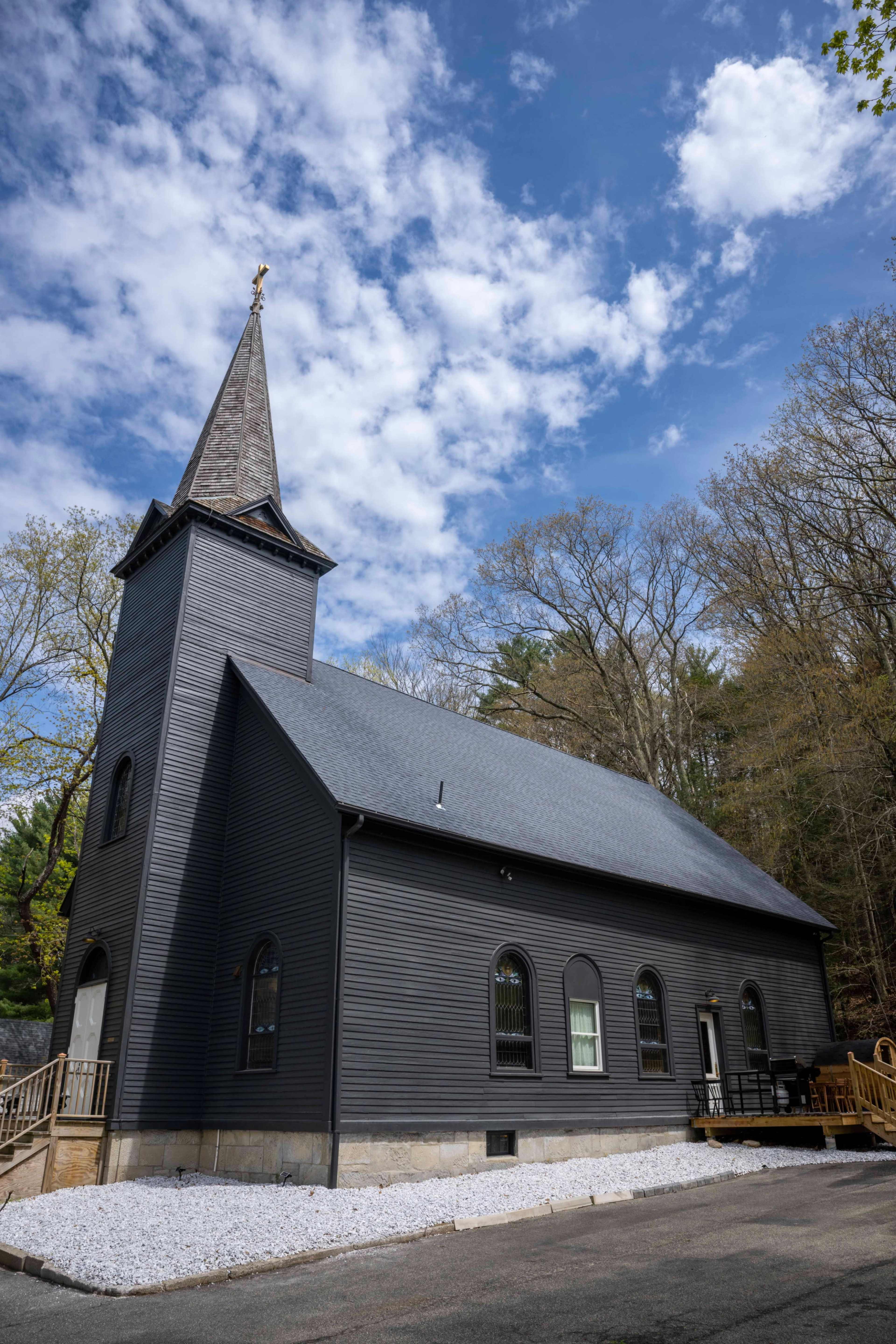A tall black church with a steeple is situated among trees under a partly cloudy sky.