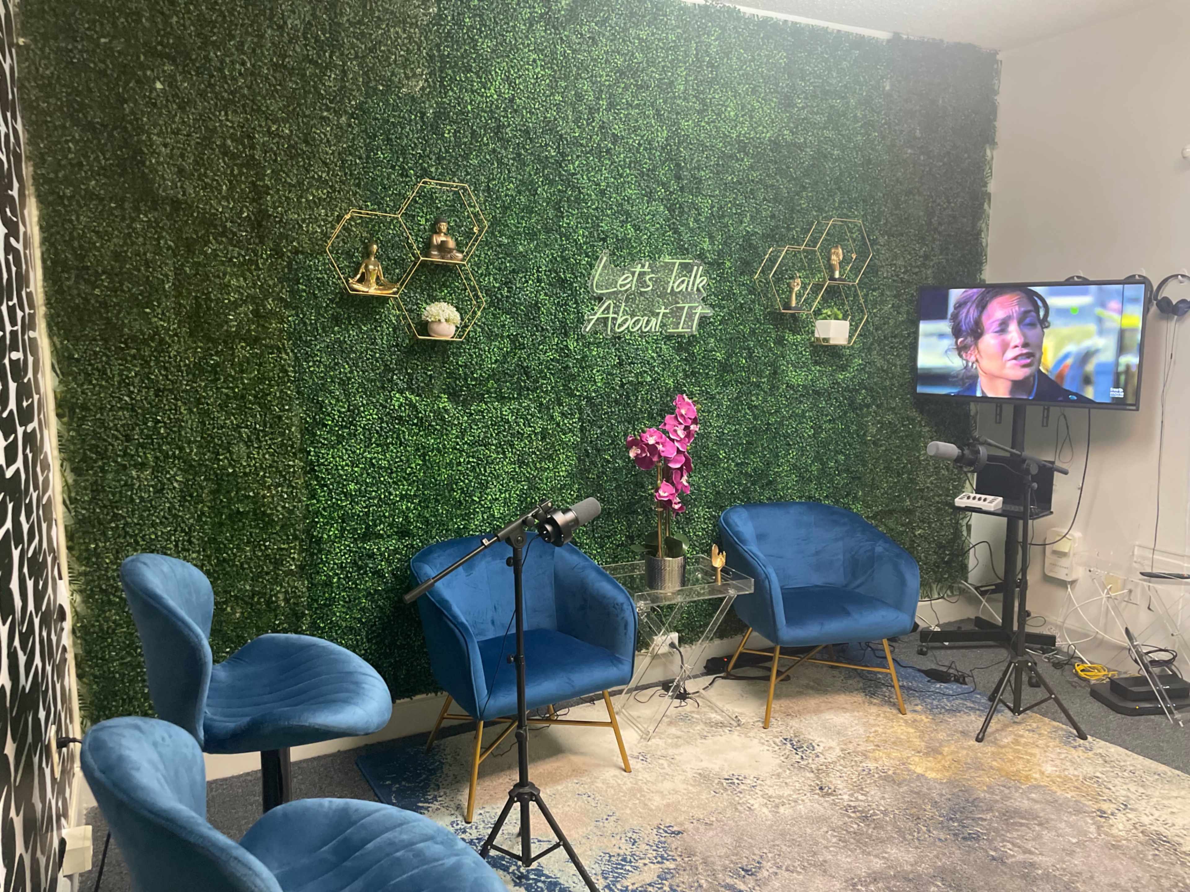 A small recording studio with three blue chairs, a decorative green wall, and a television displaying a woman speaking, alongside microphones and a plant arrangement.