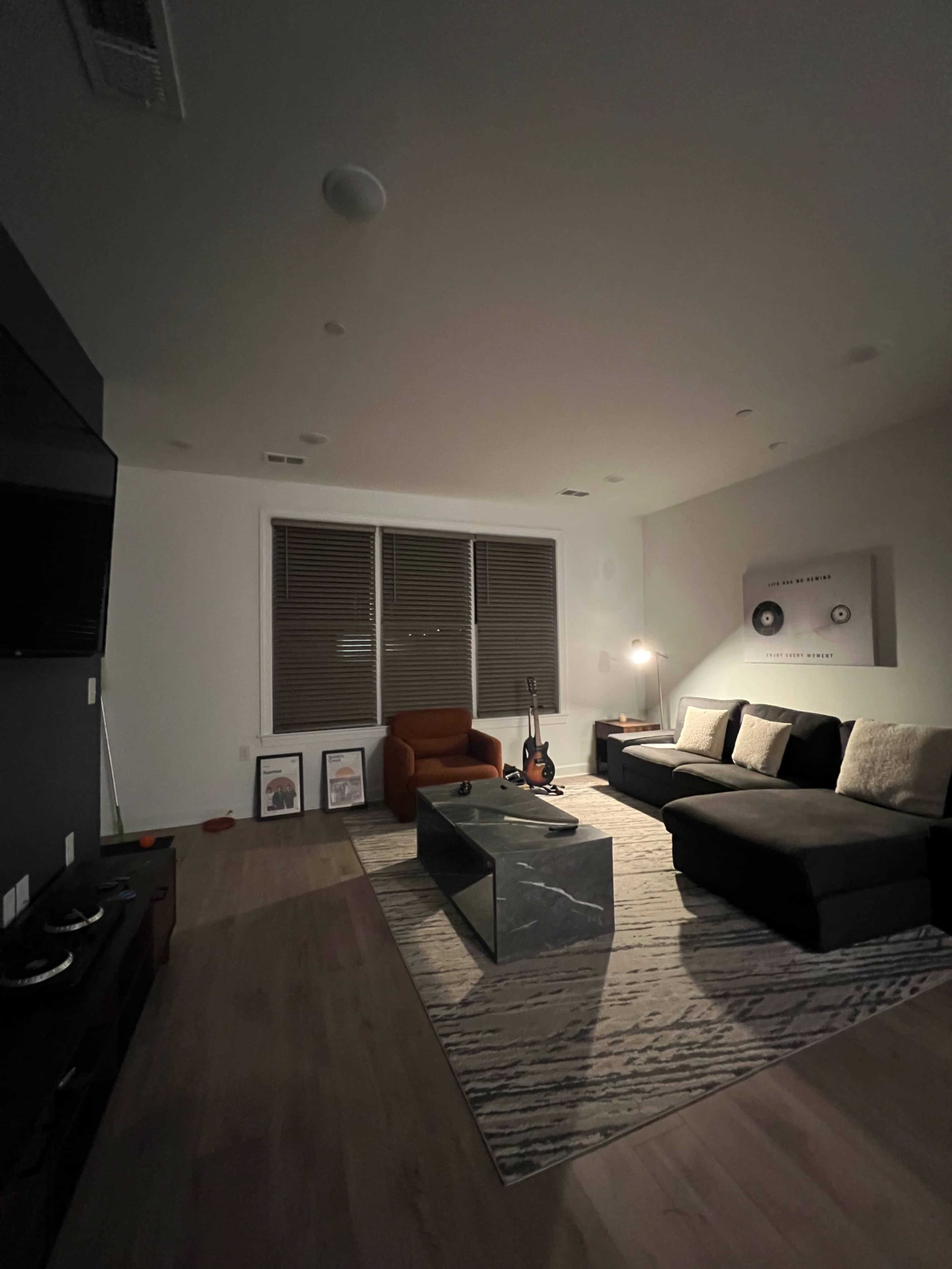 The image shows a dimly lit living room with a brown sofa, an orange armchair, a glass coffee table, and a guitar resting on the floor, alongside framed artwork on the wall.