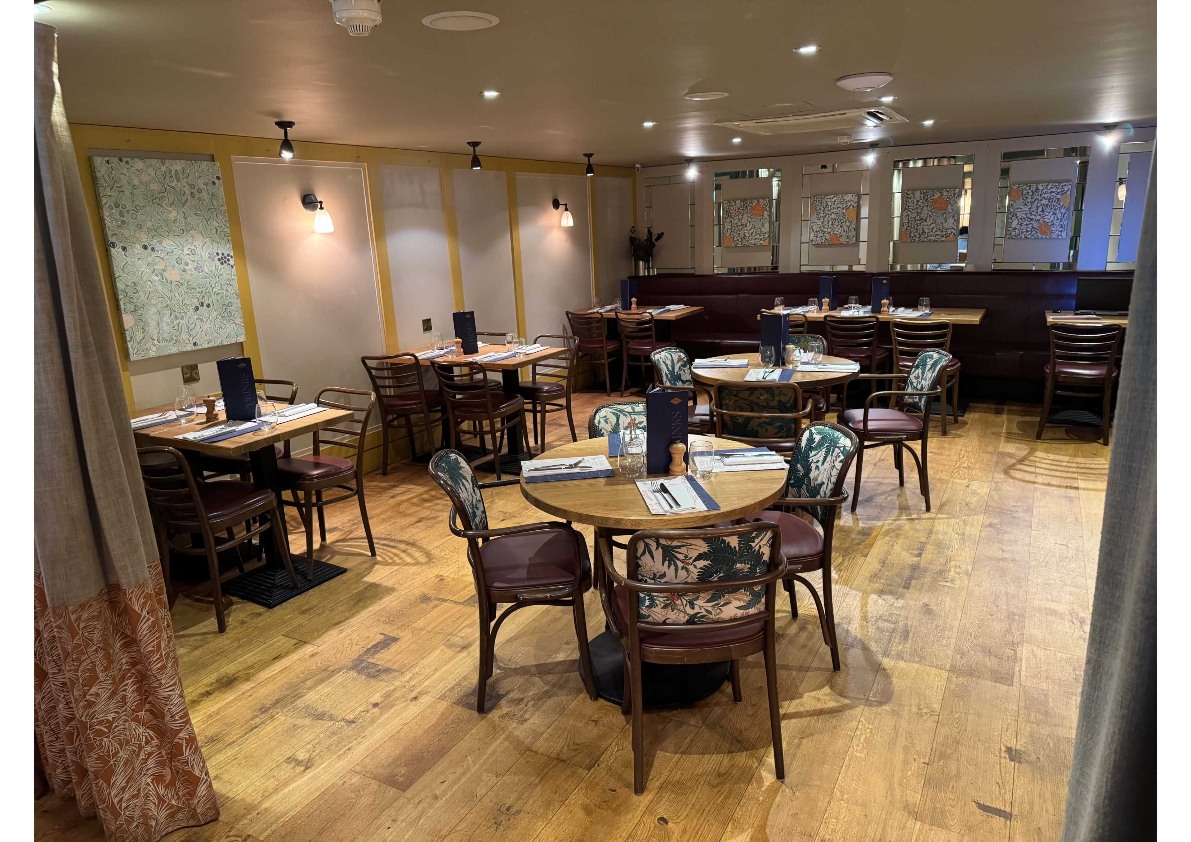 The image shows a restaurant interior with several round tables surrounded by wooden chairs, arranged on a wooden floor under soft lighting.