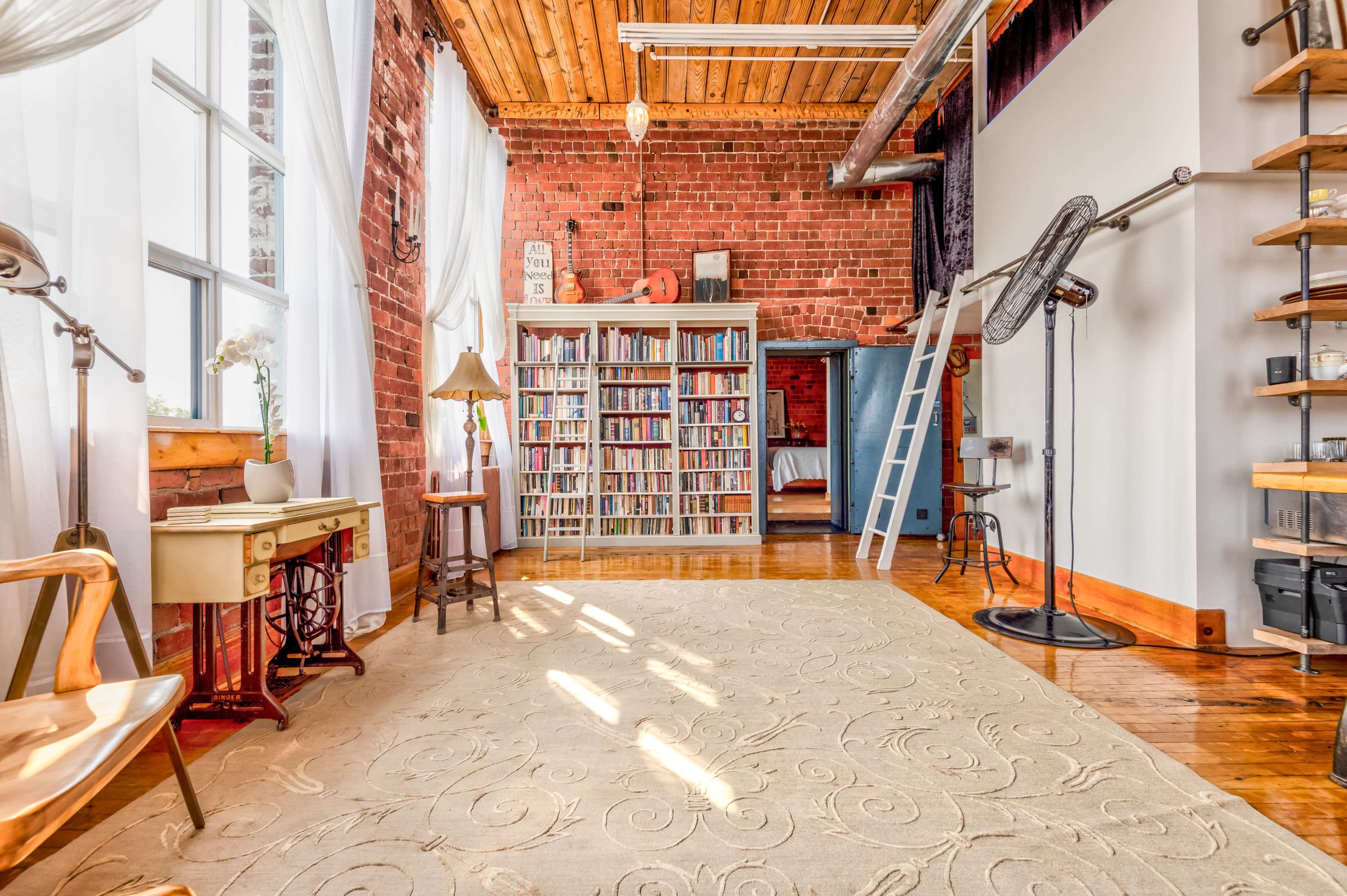 A cozy interior of a room with exposed brick walls, a large bookshelf, a light-colored rug, and wooden beams overhead.