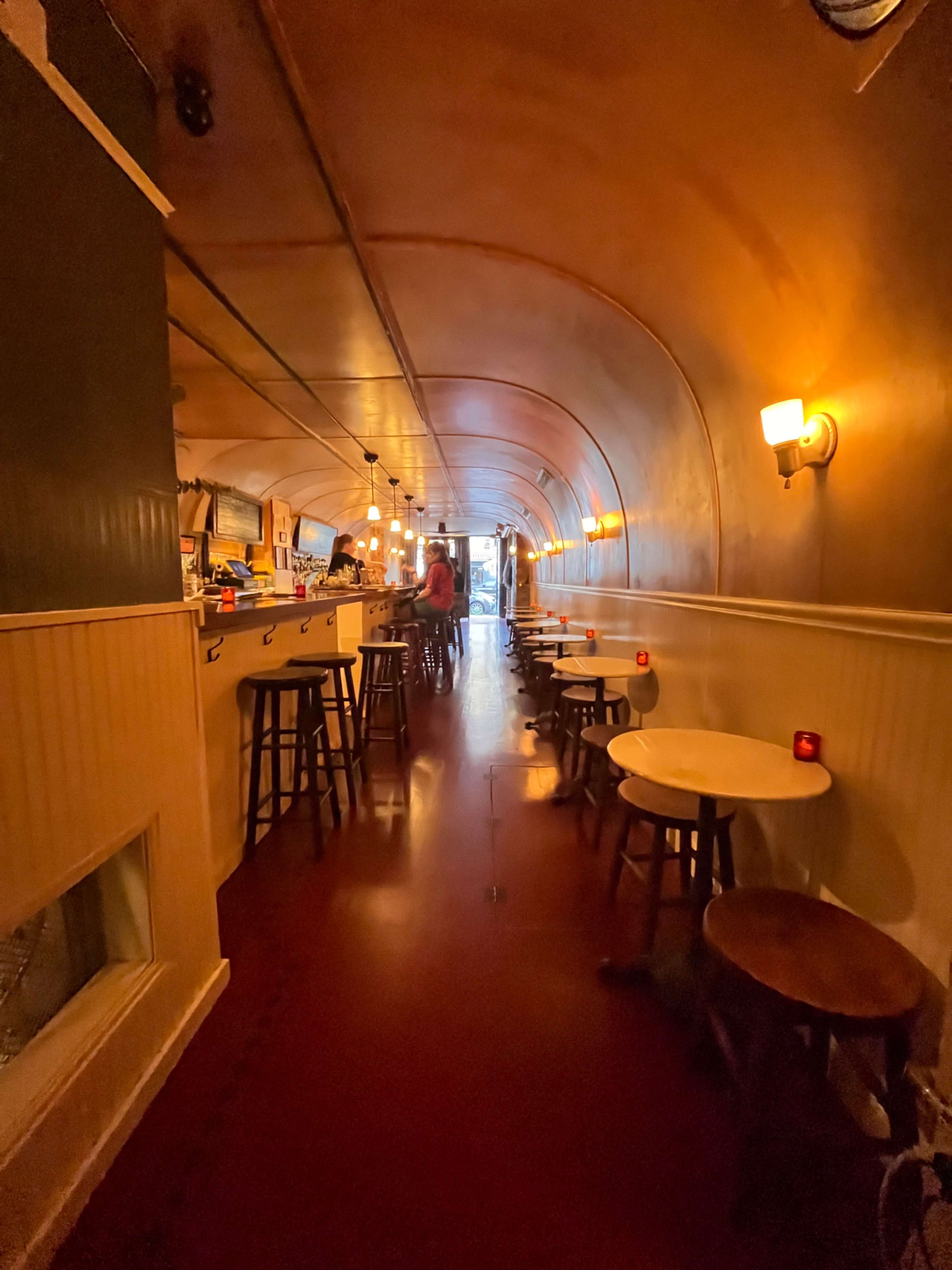 The image shows a narrow, elongated cafe interior with a curved ceiling, bar counter on one side, and small tables and stools arranged along the opposite wall.