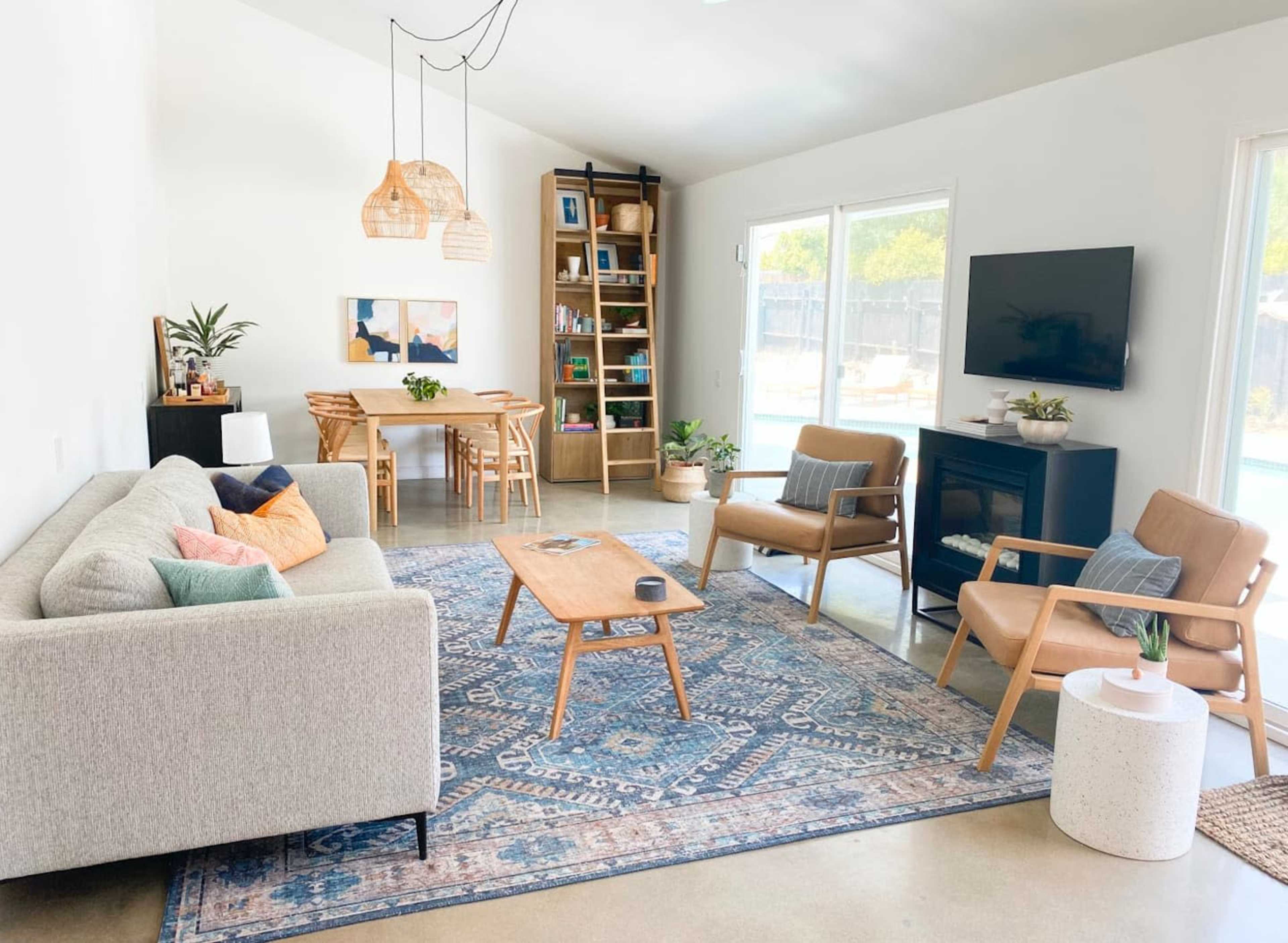 A modern living room featuring a light-colored sofa, wooden furniture, a blue patterned rug, and large windows overlooking an outdoor area.
