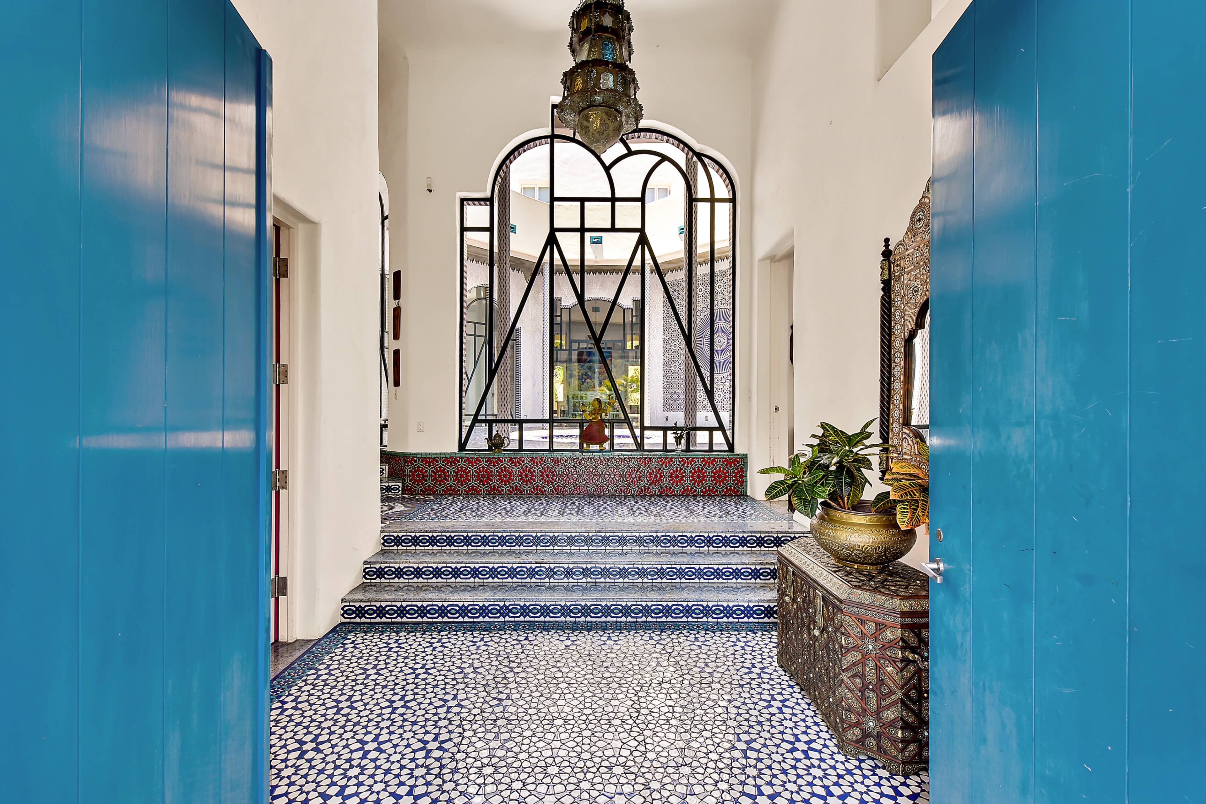The entrance hall features blue doors, intricate tile flooring, and a decorative staircase leading to a courtyard.