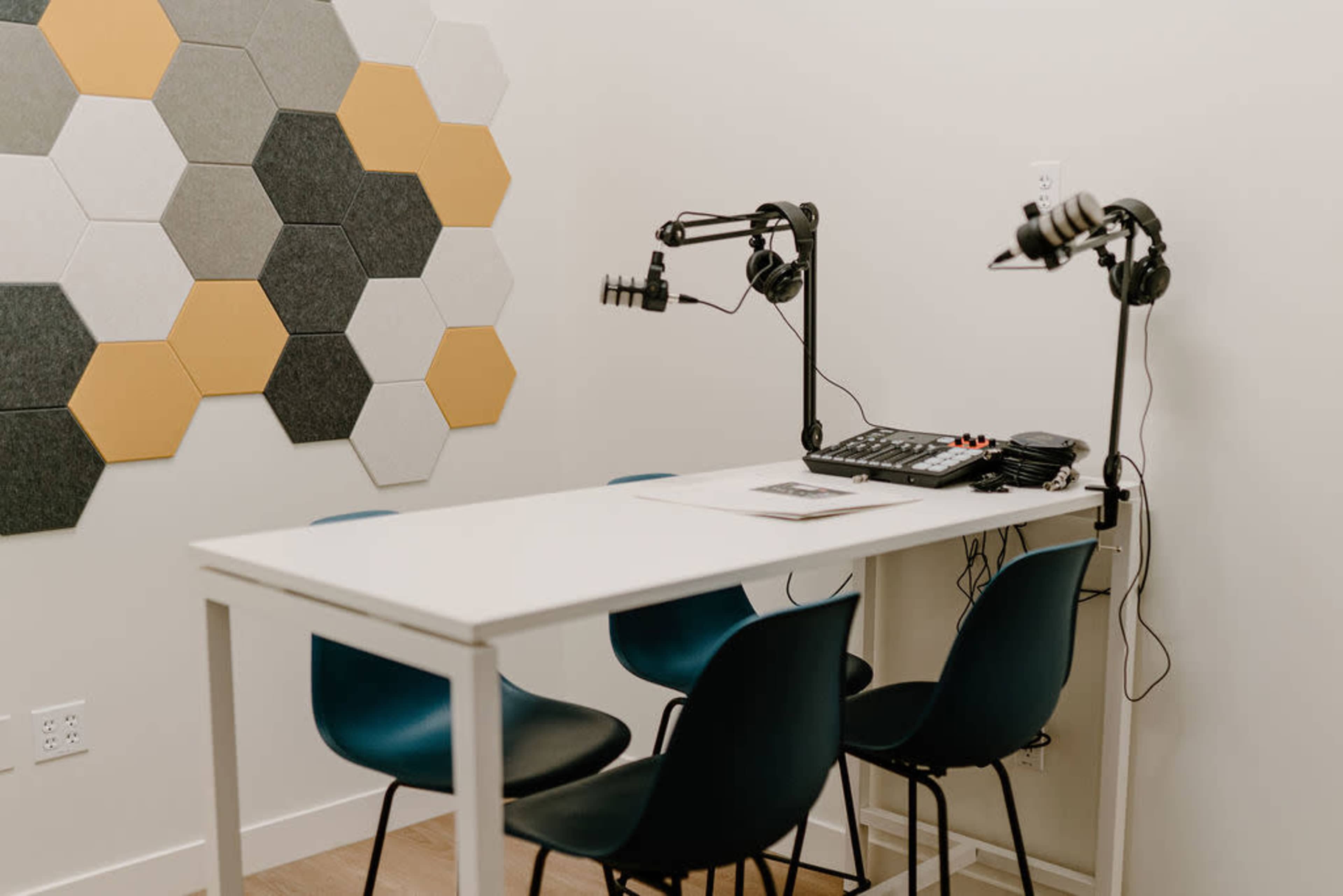A small recording studio setup featuring a white table with four blue chairs, two microphones mounted on stands, and a soundboard on the table, against a wall with hexagon-shaped acoustic panels in gray and yellow.