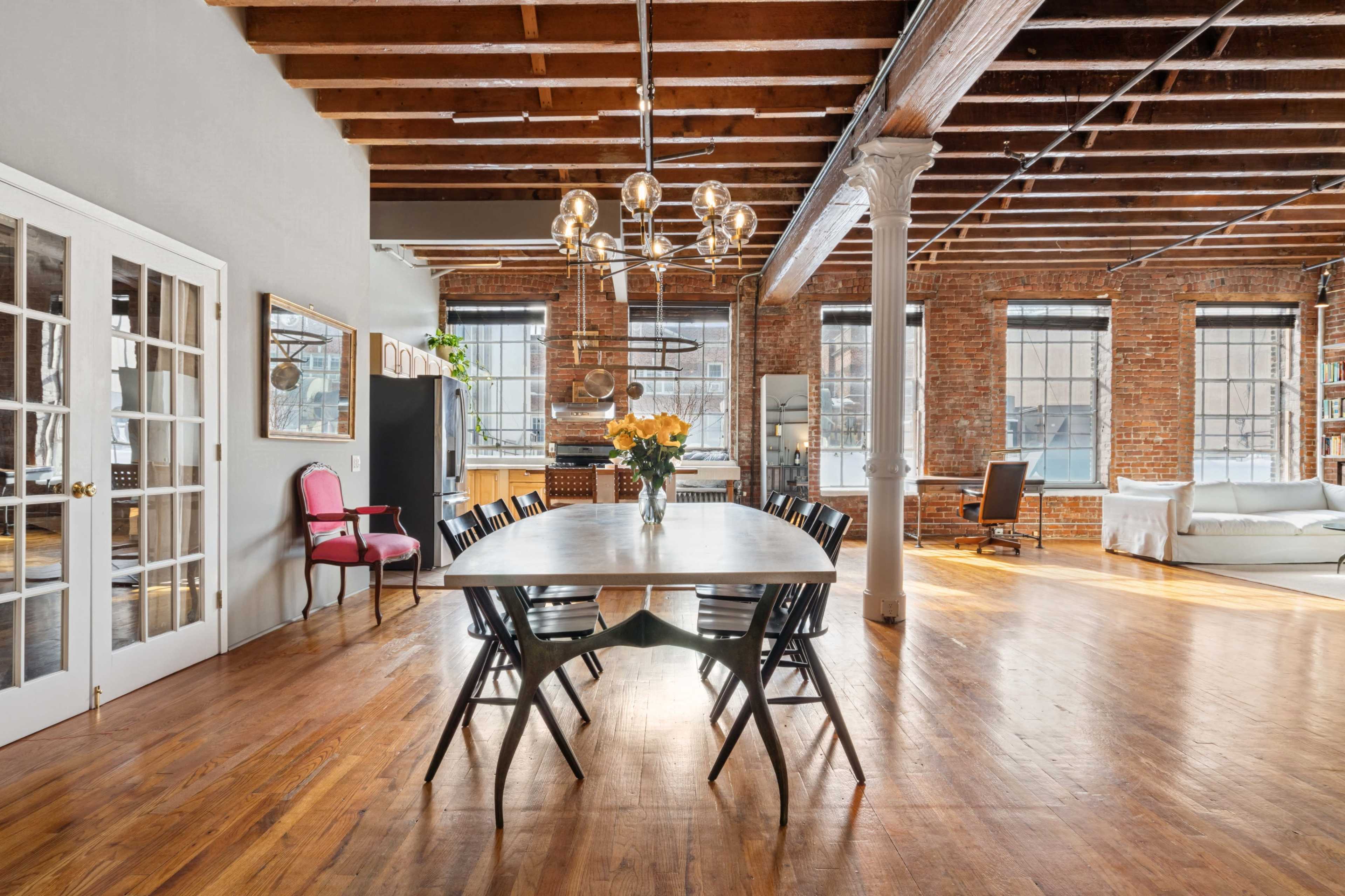 The image shows a spacious and modern loft-style living area with exposed brick walls, wooden beams, and a large dining table surrounded by chairs.
