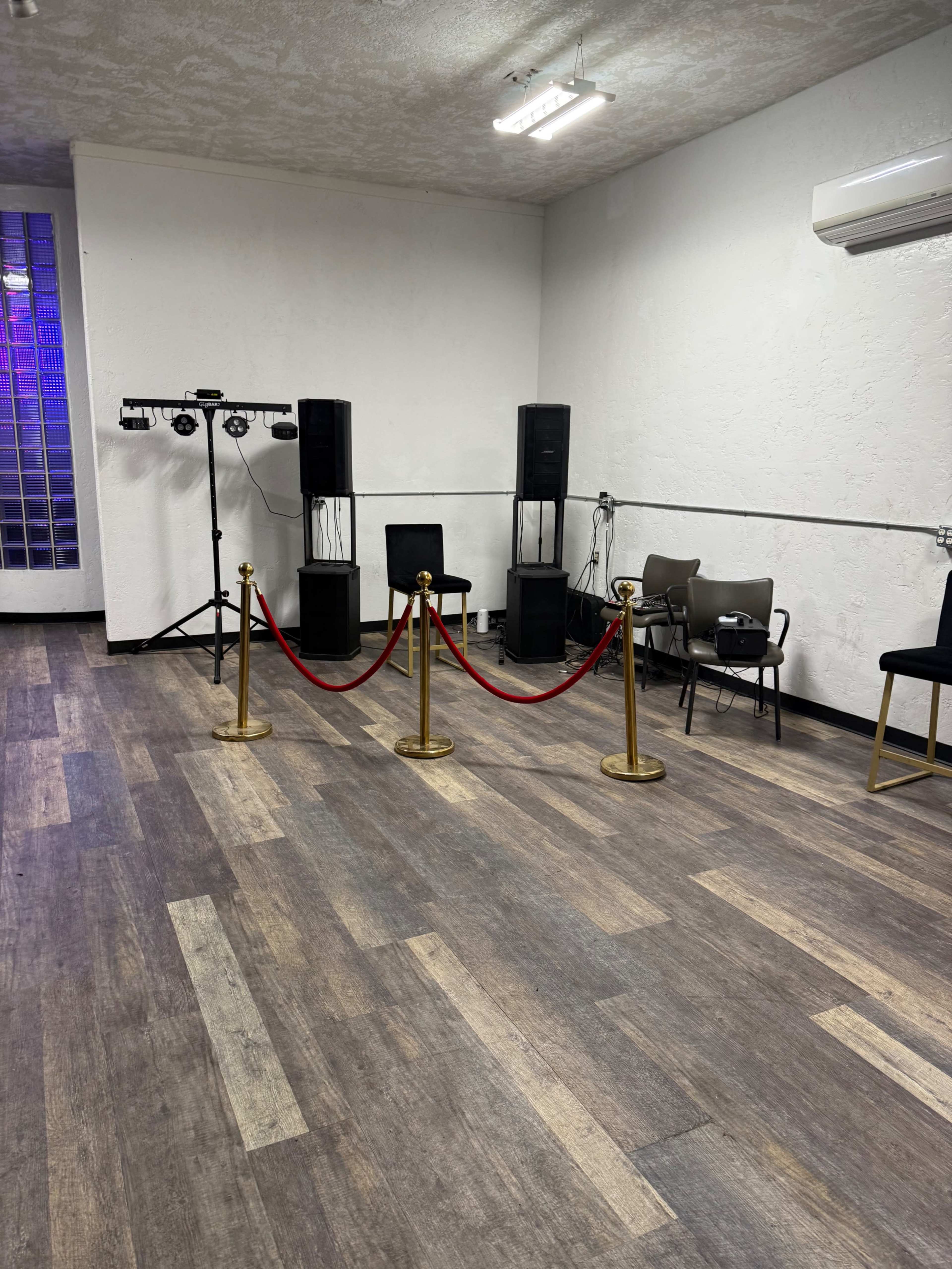 The image shows a sparsely furnished room with wooden flooring, two speakers on stands, a lighting setup, and a red rope barrier separating the area.