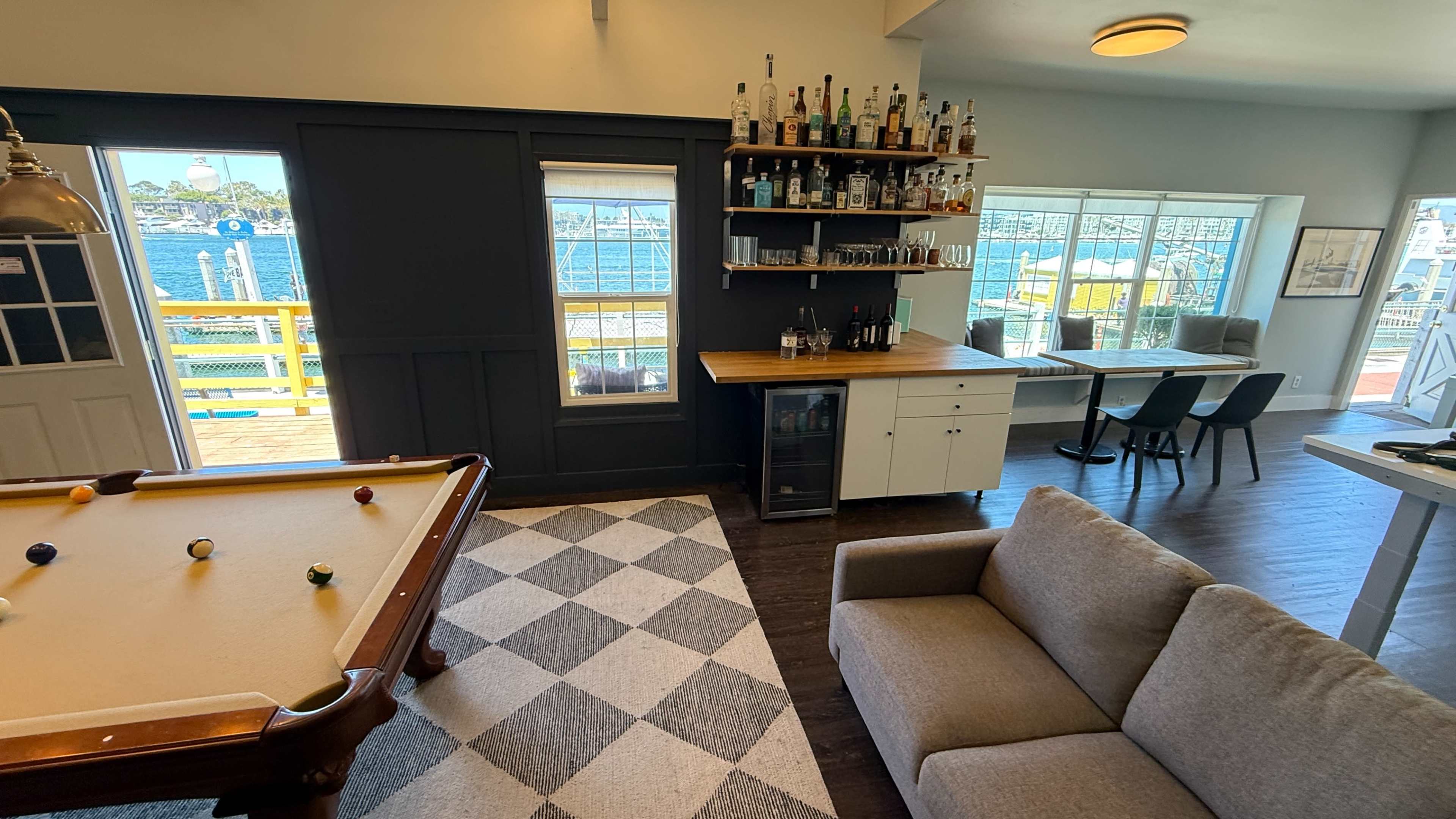 The image depicts a modern recreational room featuring a pool table, a bar area lined with bottles, a dining table, and large windows overlooking a waterfront view.