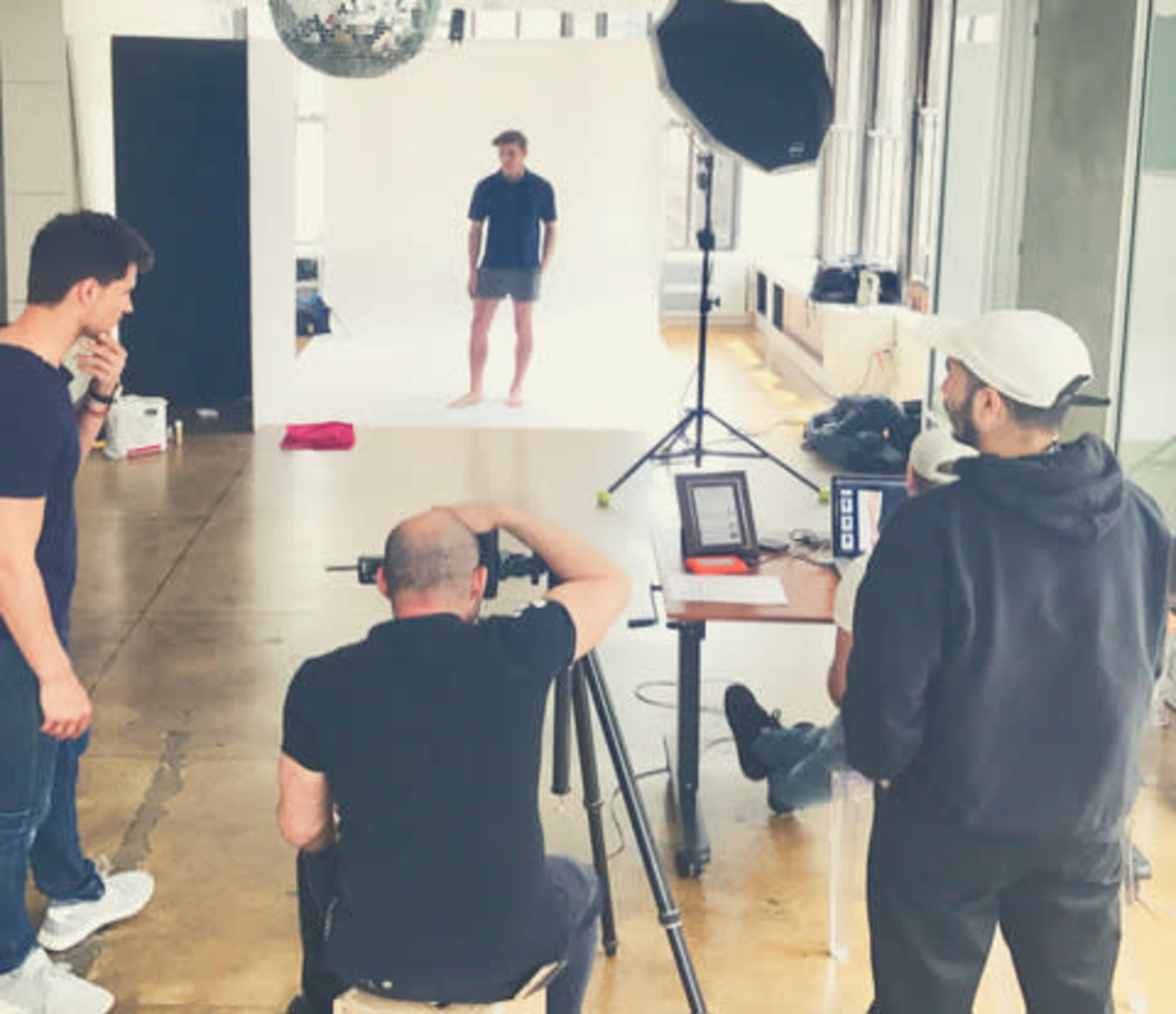 A group of four people is gathered in a photography studio, with one person posing against a white backdrop while others operate equipment and review images.