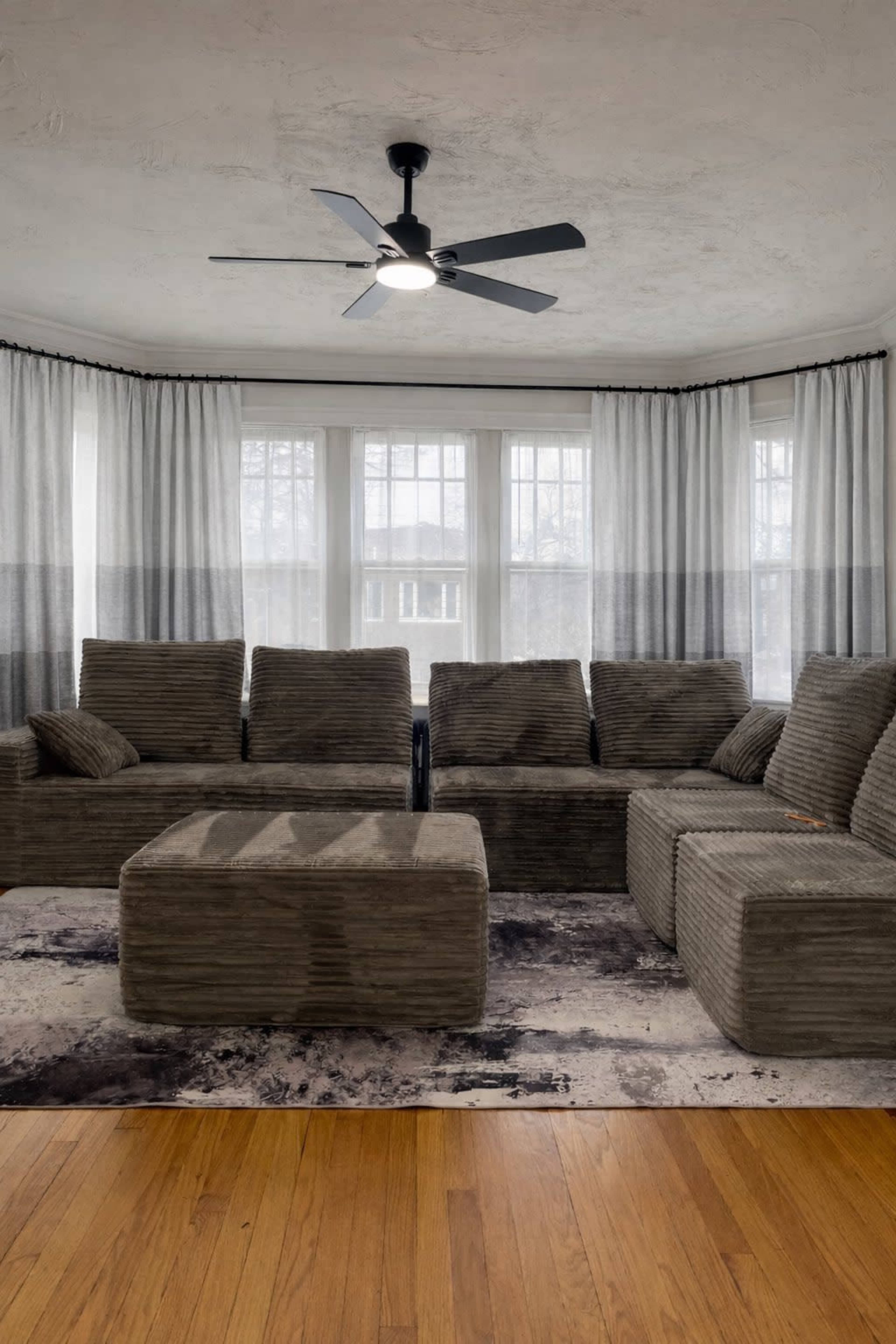 The image shows a living room featuring a sectional sofa, an ottoman, and large windows covered with sheer curtains, all positioned on a hardwood floor.